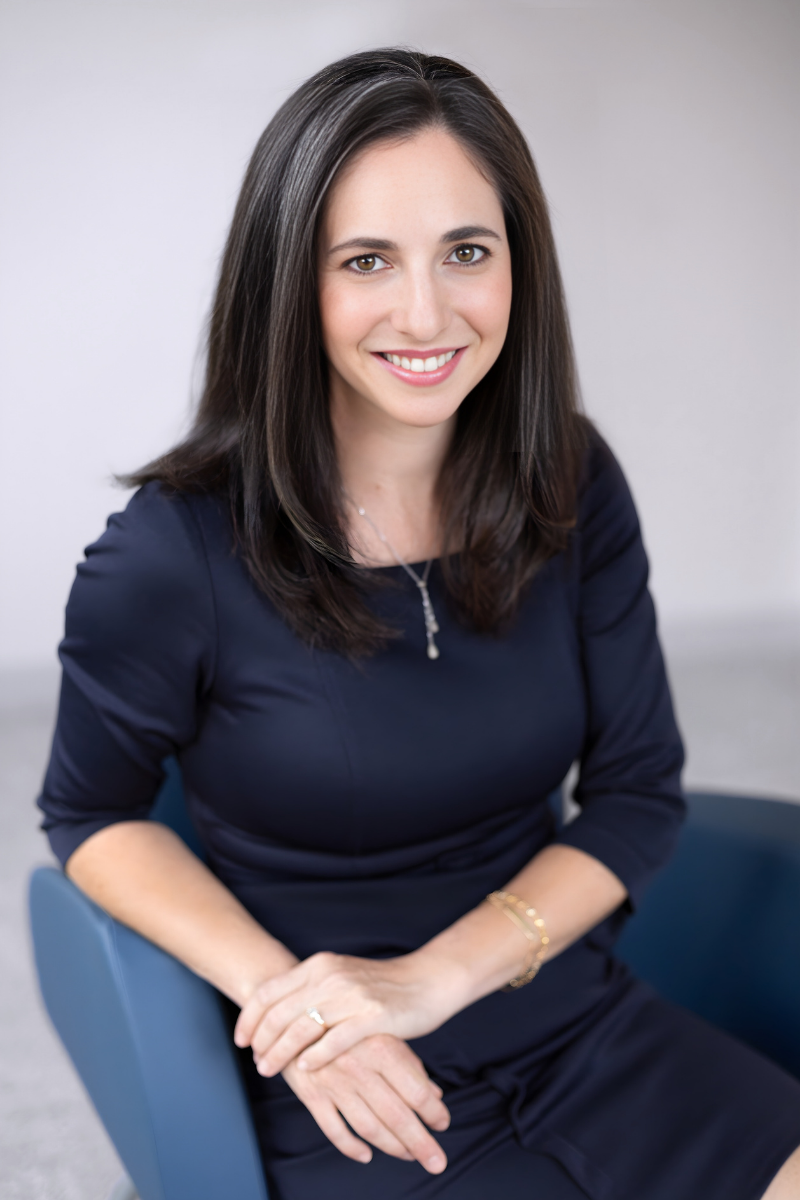 A woman sitting on a gray couch in a modern indoor space with green wall and wooden accents, wearing a white long-sleeve shirt and blue jeans, smiling at the camera.