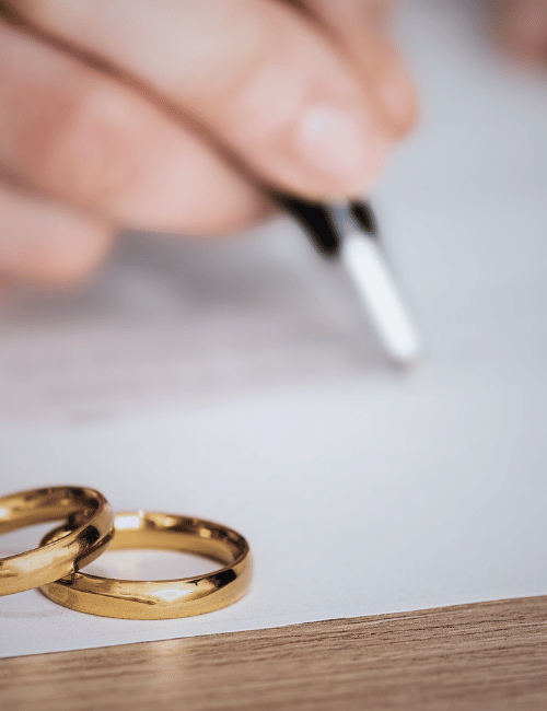 Close-up of two gold wedding rings on a table, with a hand in the background holding a pen, possibly signing a document.