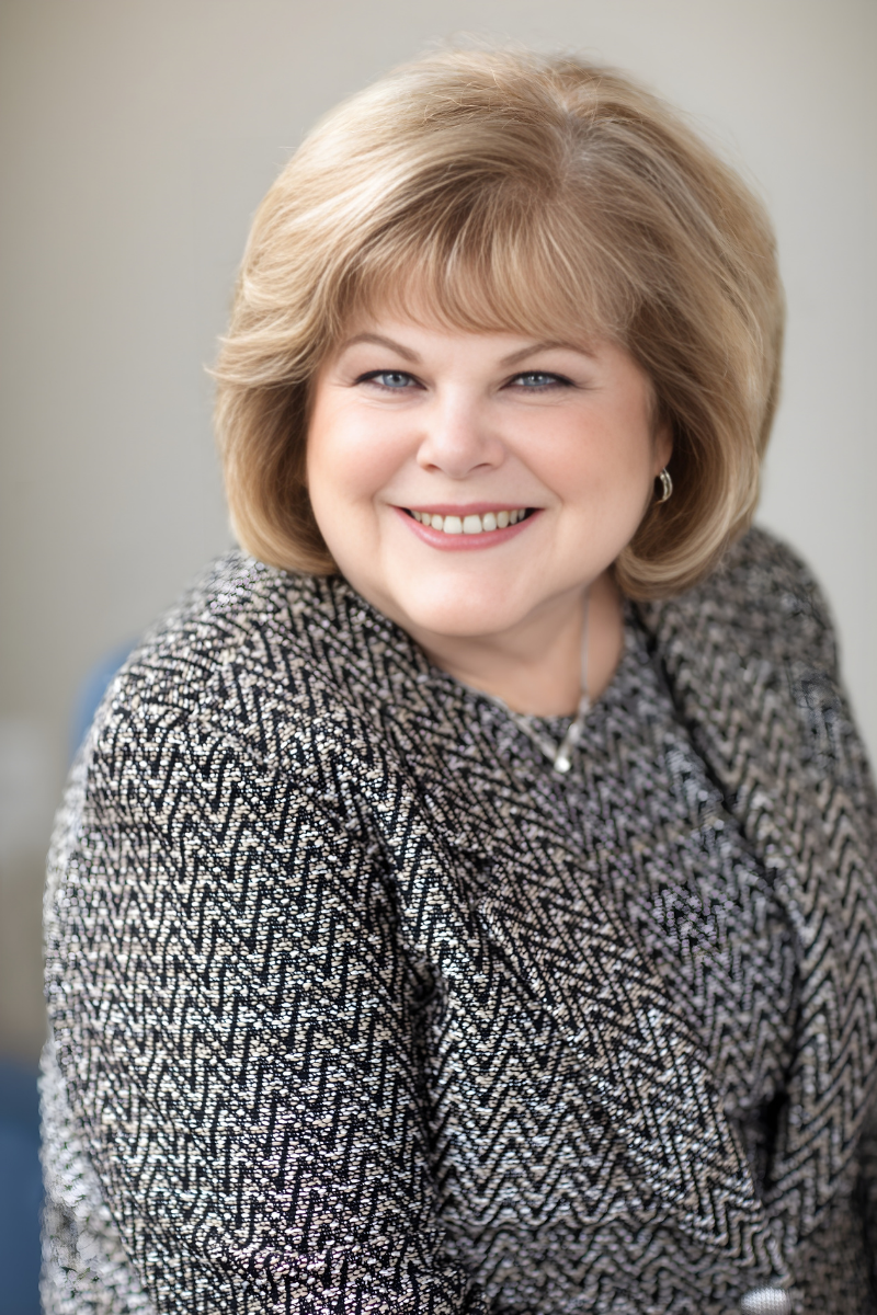 A smiling middle-aged woman with blonde hair, wearing a black top and a colorful scarf, resting her chin on her clasped hands, showing rings and a bracelet.