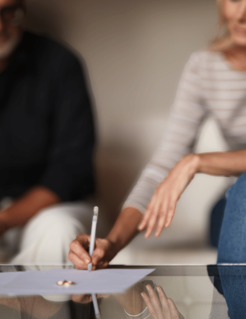 A woman signing documents on a table during a meeting or legal process with two other people present.