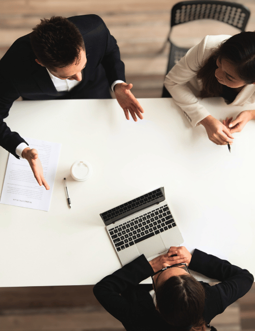 Three people having a business meeting at a white table, discussing documents and a laptop.