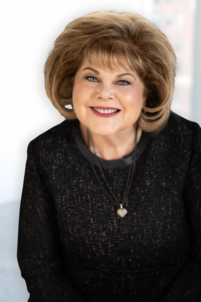 A smiling middle-aged woman with blonde hair, wearing a black top and a colorful scarf, resting her chin on her clasped hands, showing rings and a bracelet.