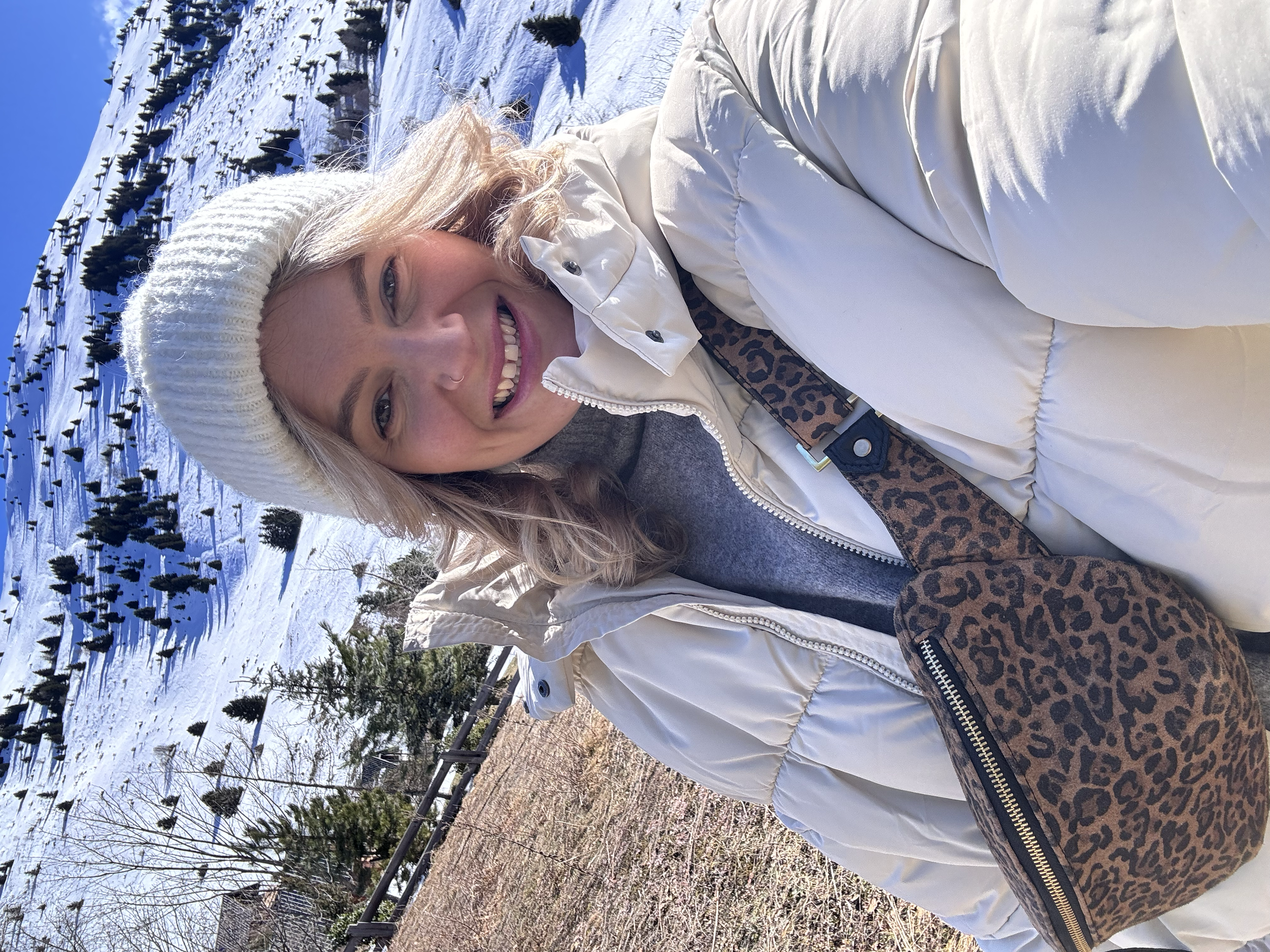 Holistic coach, loose weight without restriction. premium nutrition coaching. Woman smiling outdoors in a snowy landscape, wearing a white beanie, white puffer jacket, and a leopard print crossbody bag.