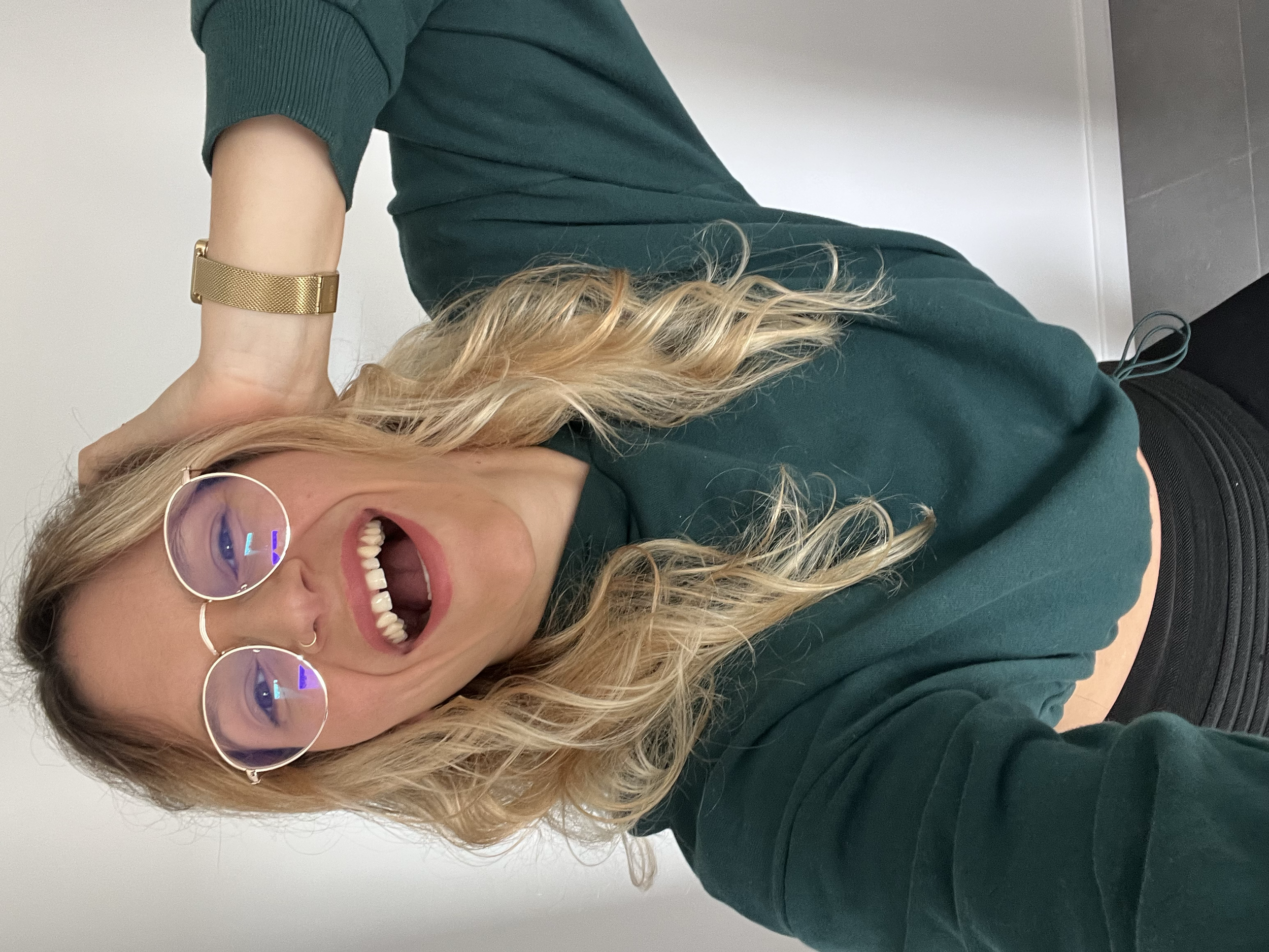 Holistic coach, loose weight without restriction. premium nutrition coaching. Smiling woman wearing sunglasses with purple reflection, green sweater, black pants, and a watch, posing indoors against a plain white wall.