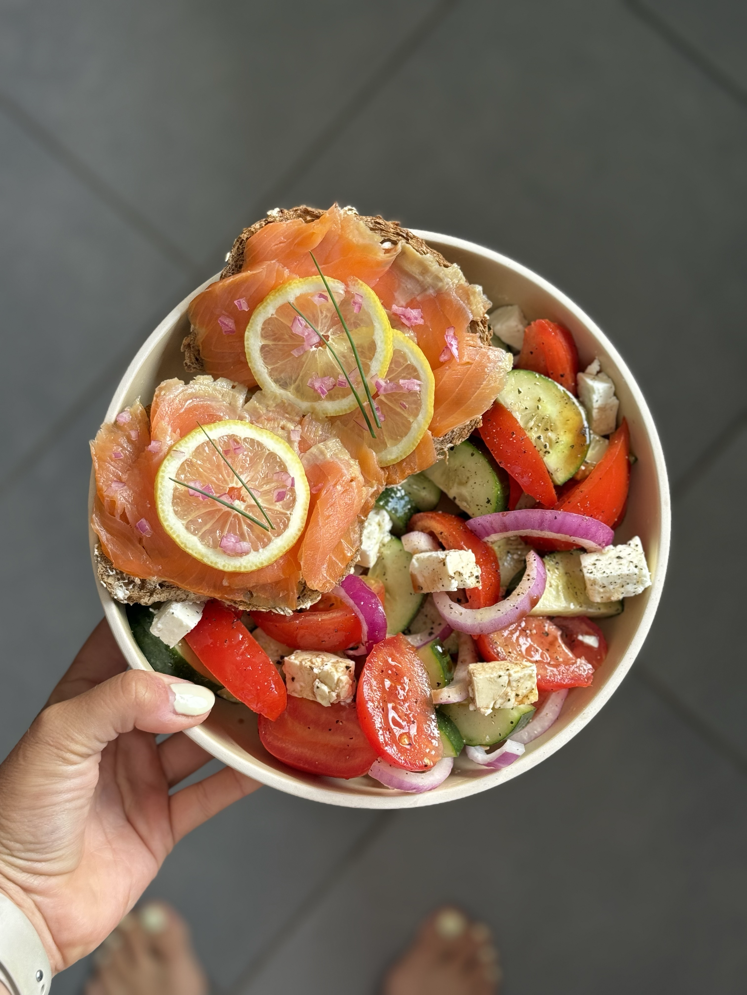 A bowl of salad containing sliced cucumbers, tomatoes, red onions, tofu, and sliced lemon on top of smoked salmon on bread slices garnished with chives and pink pepper flakes.
