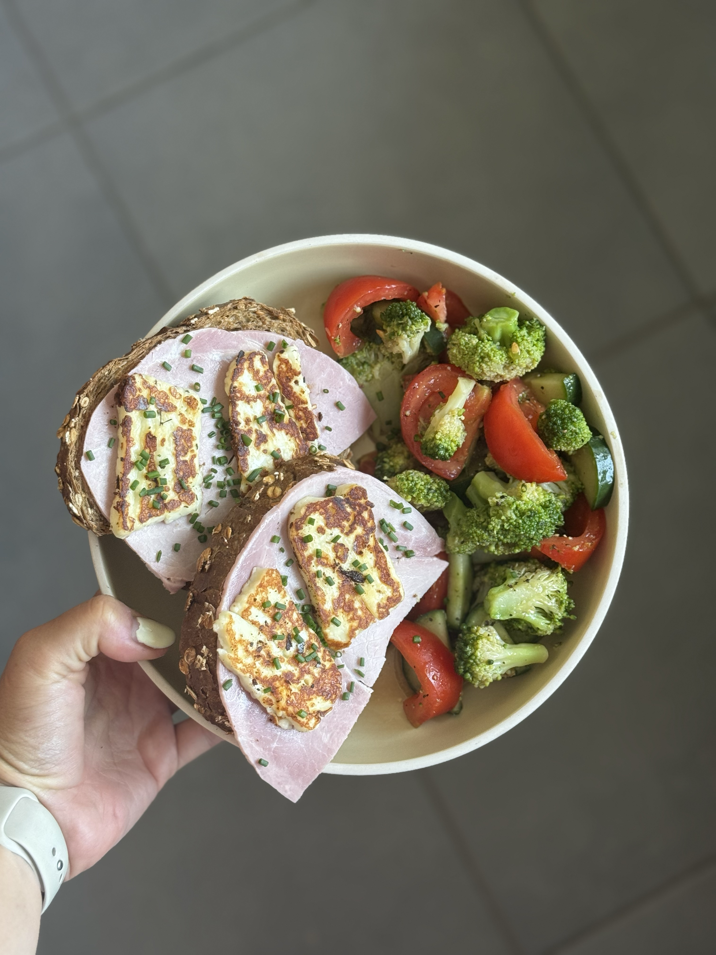 Hand holding a bowl with sliced ham and grilled cheese sandwiches on multigrain bread, alongside a salad of broccoli, cherry tomatoes, and cucumber.