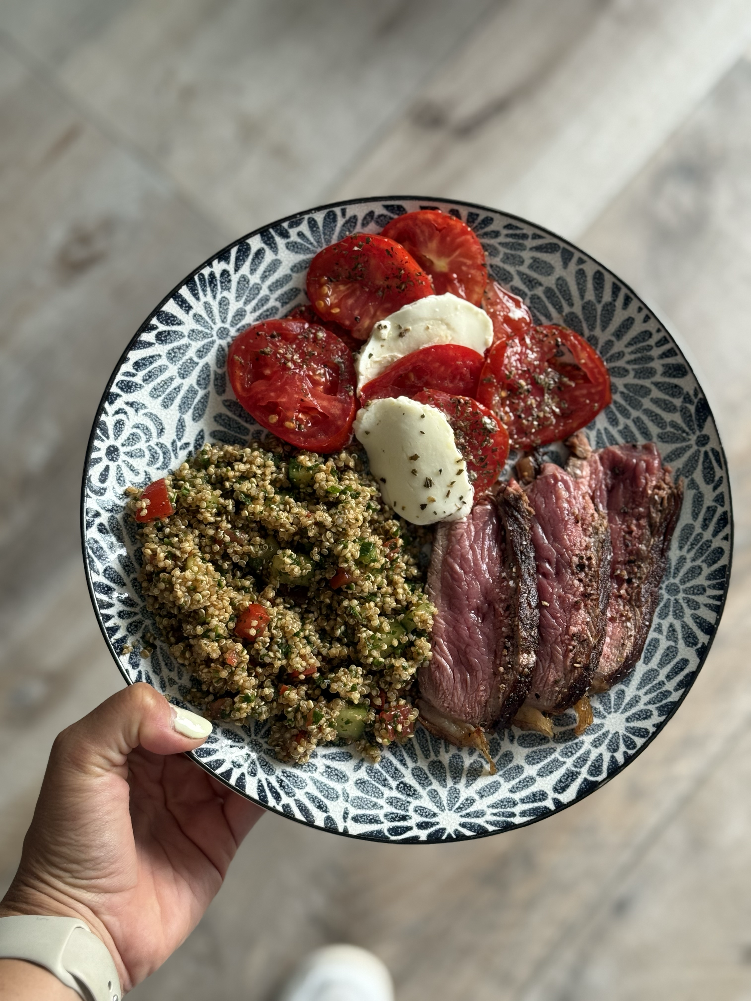 A plate with sliced tomatoes, mozzarella cheese, cooked steak, and quinoa salad, held in a person's hand over a wooden floor.