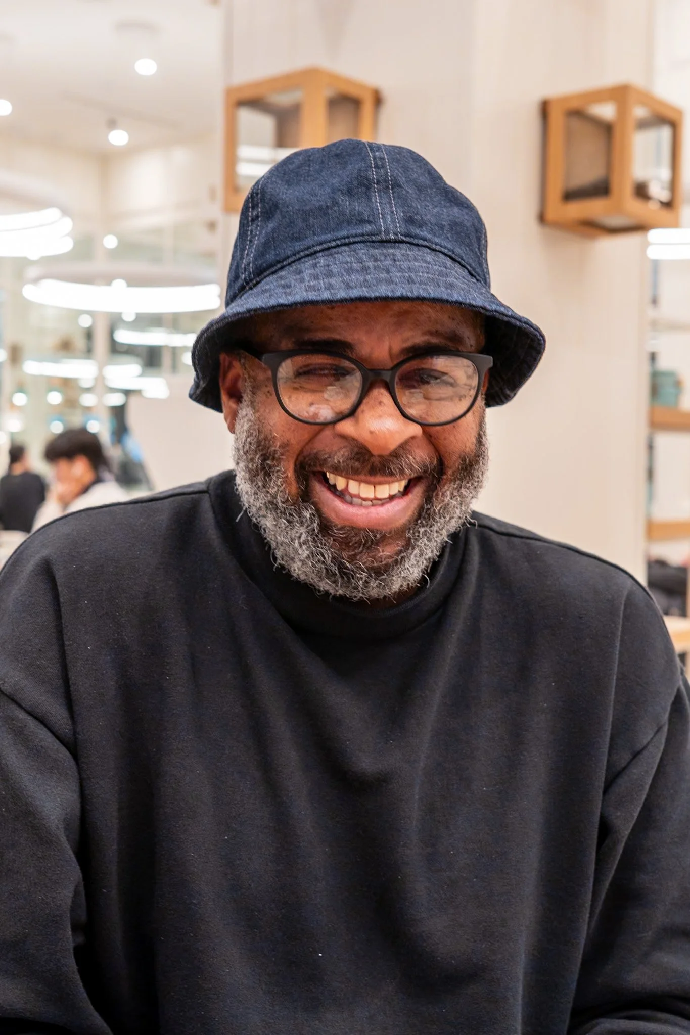 A smiling man with glasses, a beard, and a denim bucket hat, sitting indoors in a well-lit environment.