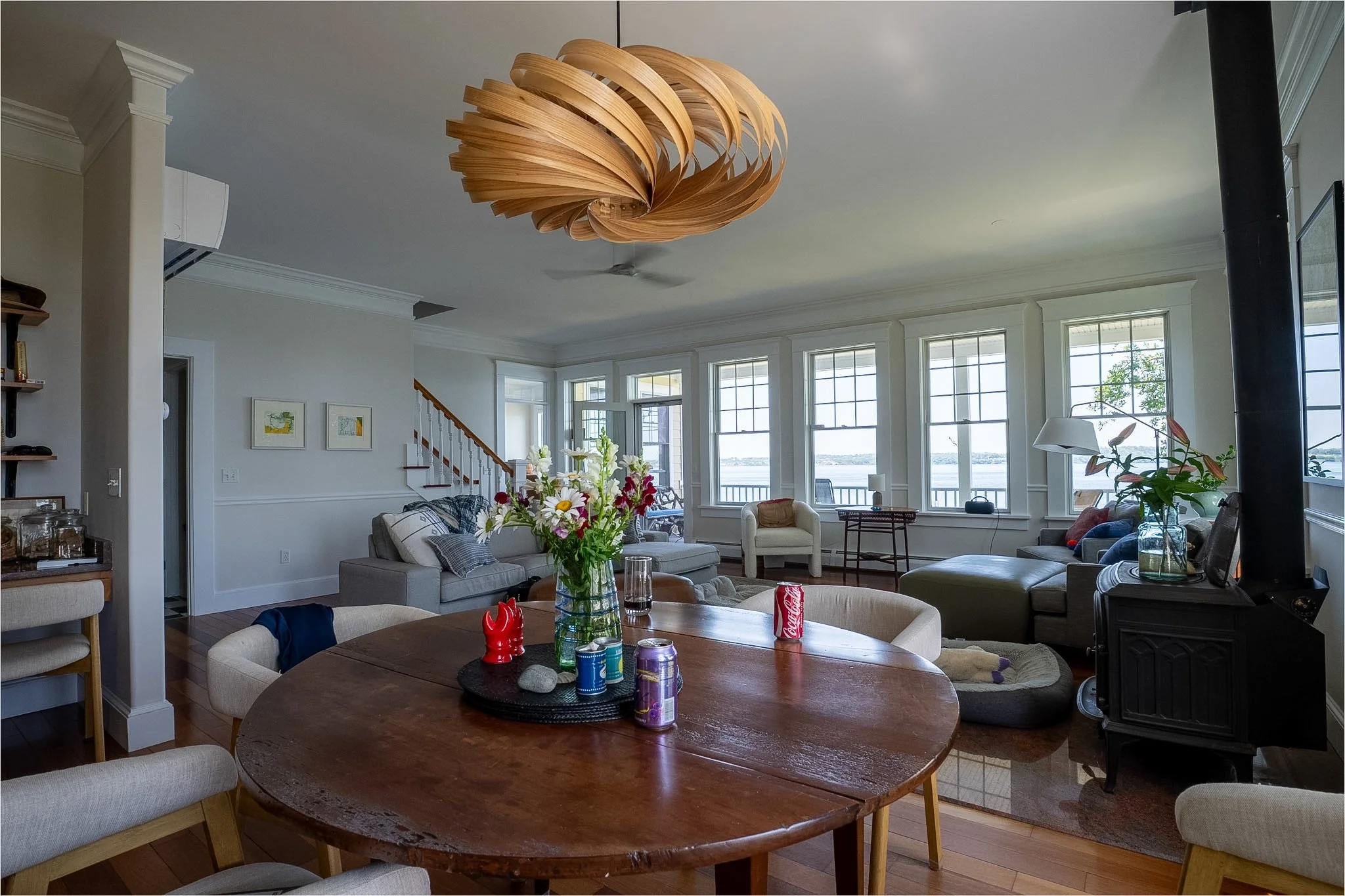 A living room with a wooden dining table, upholstered chairs, a gray sofa, a floral centerpiece, and a chandelier hanging from the ceiling. Large windows show a view outside. Various decorative items and artwork are on the walls.