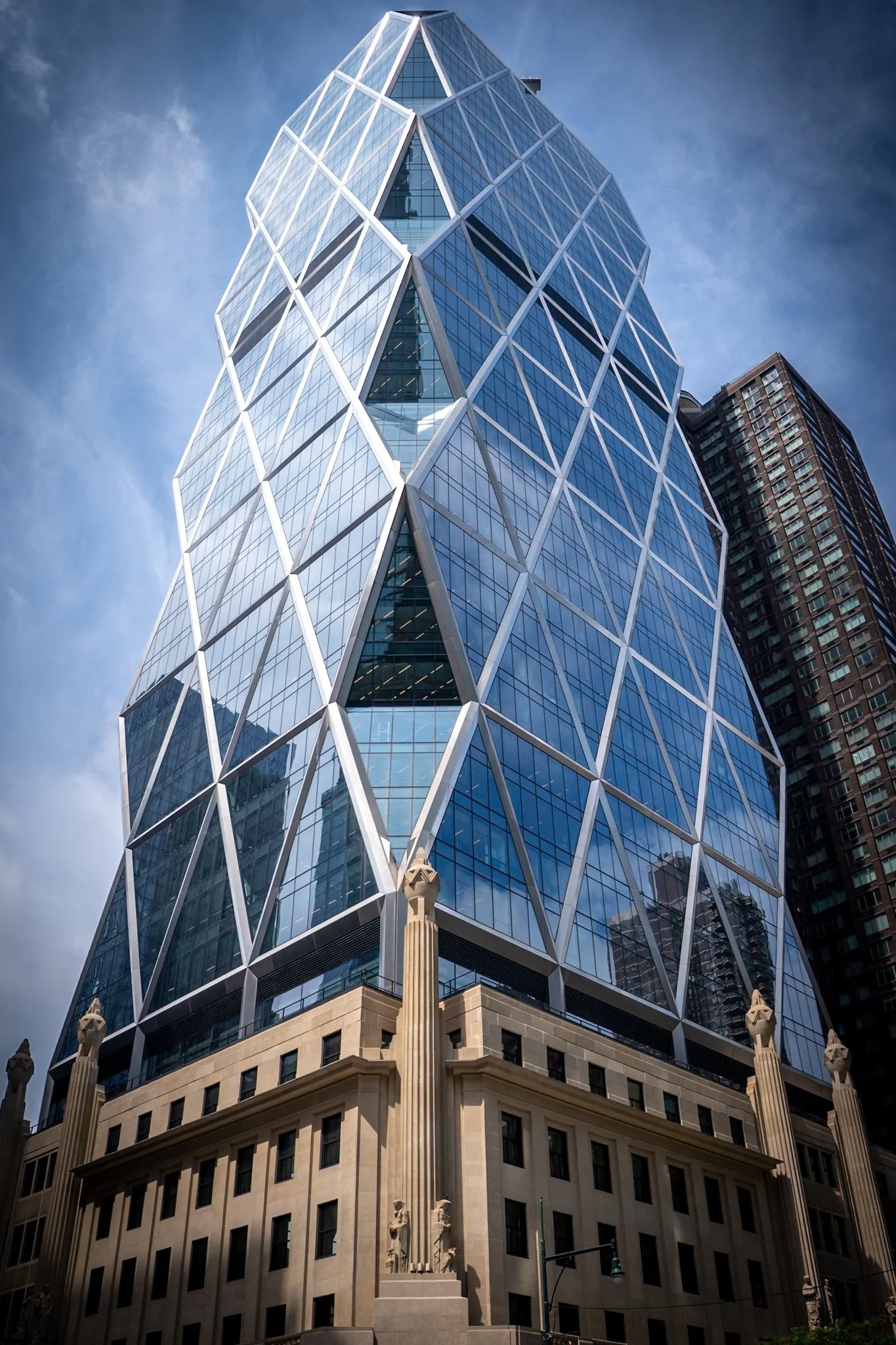 A modern skyscraper with a glass exterior featuring a geometric design, standing above a historic building with classical architecture and decorative sculptures at street level.