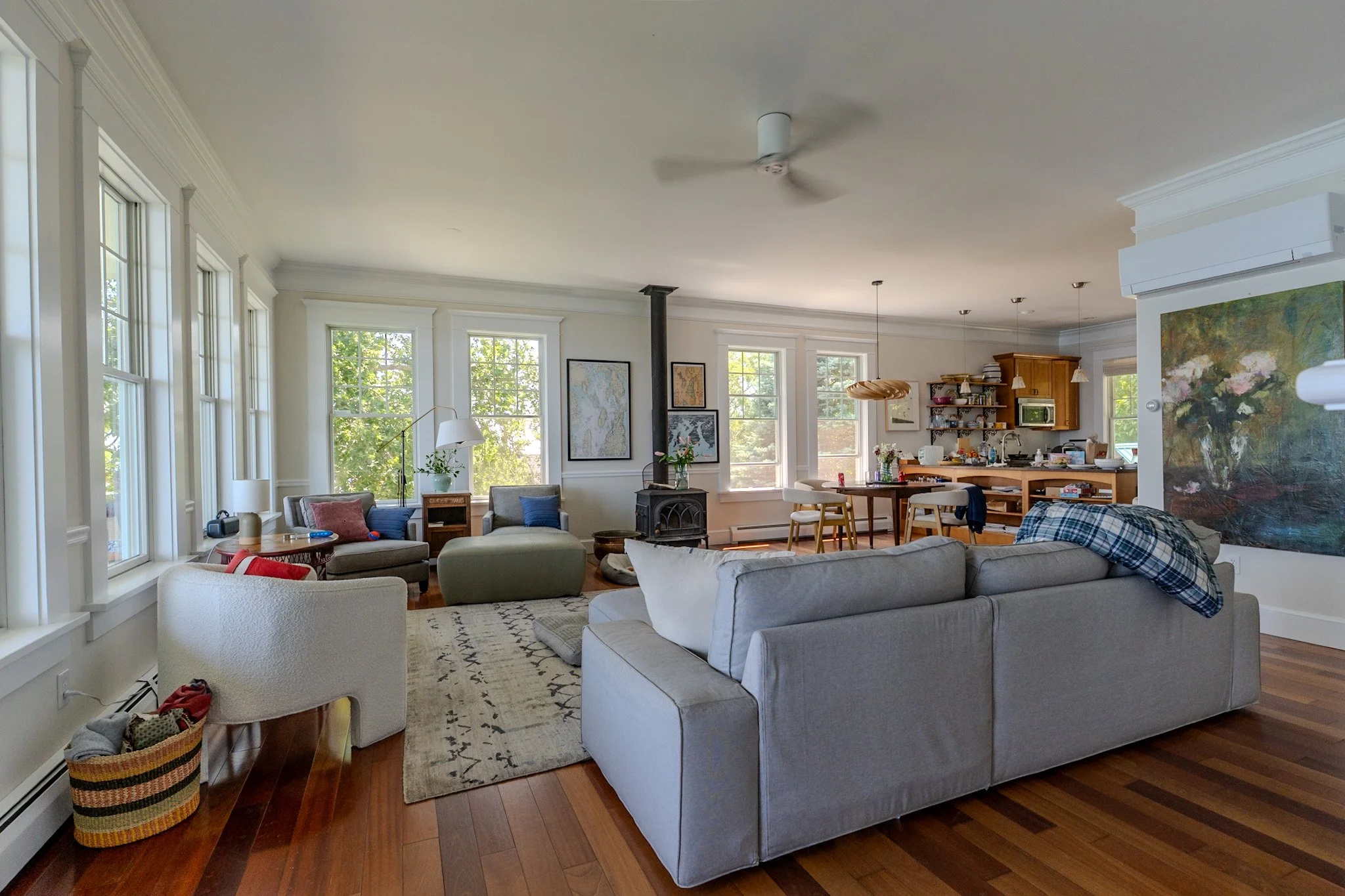 A living room with large windows, a white sofa, armchair, area rug, and wooden flooring. The kitchen area is visible in the background with wooden cabinets, open shelves, and a dining table with chairs. There are paintings on the walls and various de