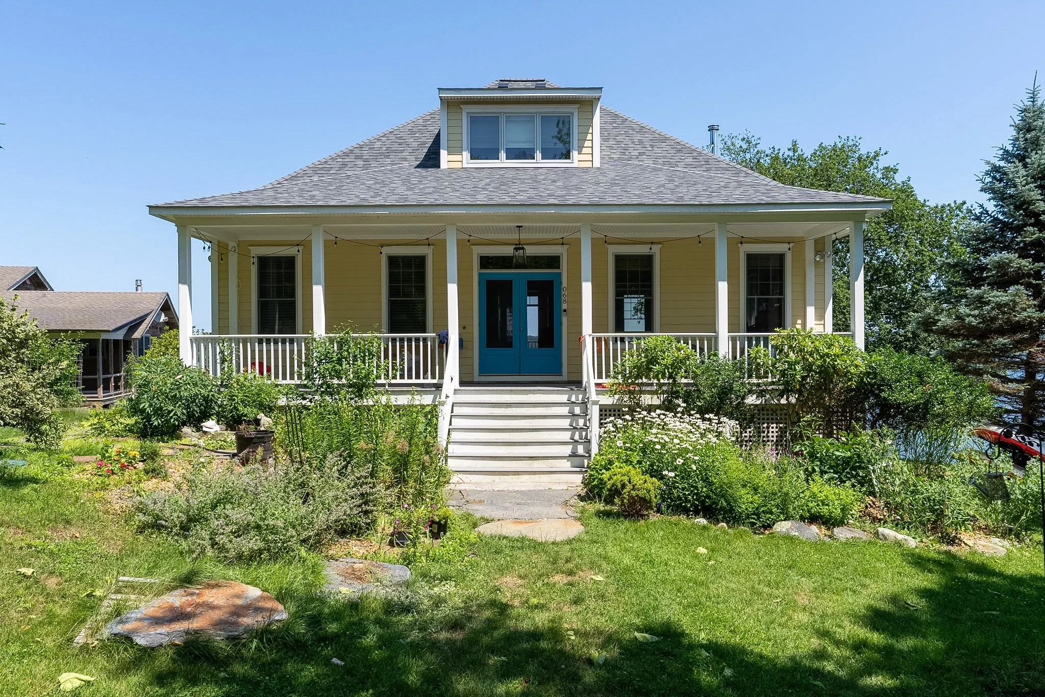 Yellow house with white porch, blue front door, stairs, garden, trees, and clear blue sky.