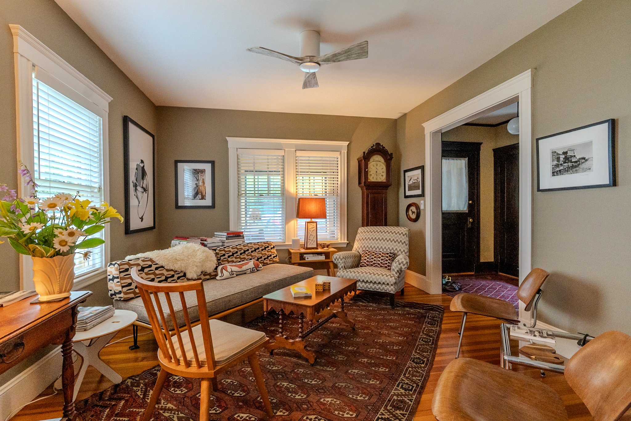 Living room with green walls, white trim, and hardwood floors. Furniture includes a patterned sofa, a wooden dining table with chairs, a light-colored armchair, and a side table with a lamp. Decor features framed artwork on the walls, a tall grandfat