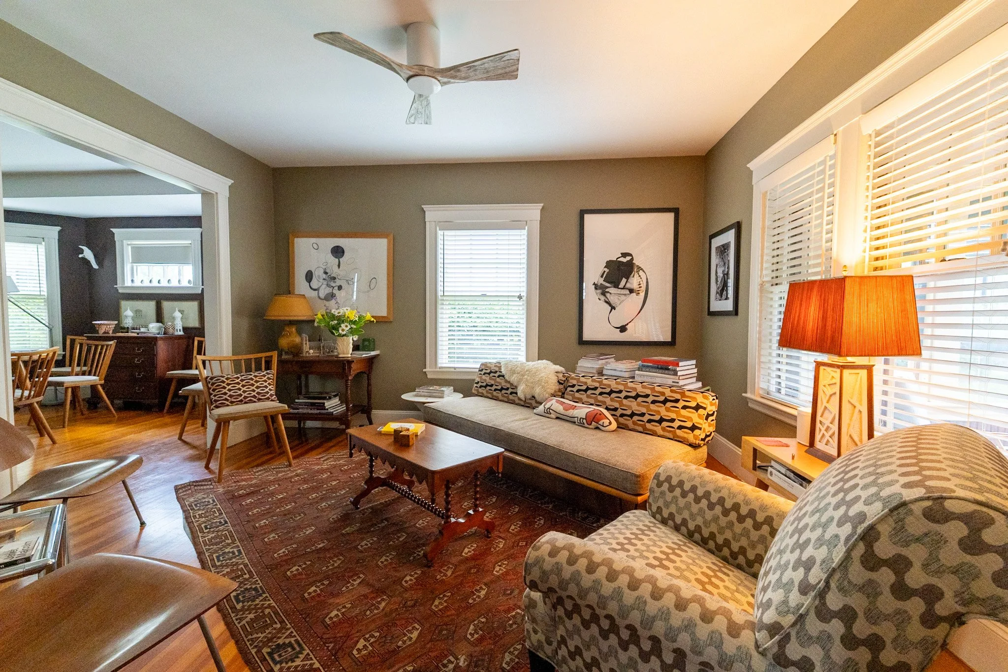 A cozy living room with beige walls, hardwood floors, a large window with white blinds, and various framed artworks. There is a patterned sofa with pillows, a wooden coffee table, a side table with a yellow lamp, and a stack of books. The room is wel