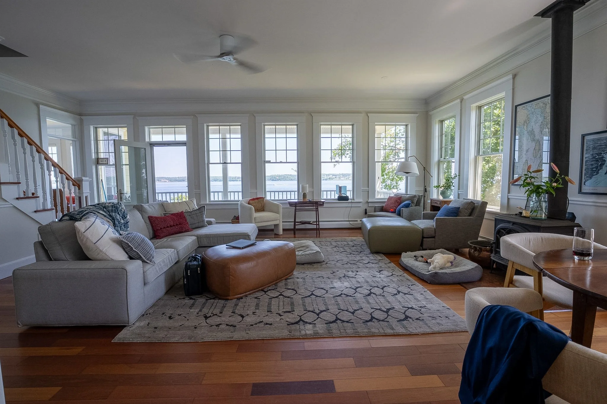 Living room with large windows overlooking water, white sofas with colorful pillows, a television, a cozy rug, and wooden floors.