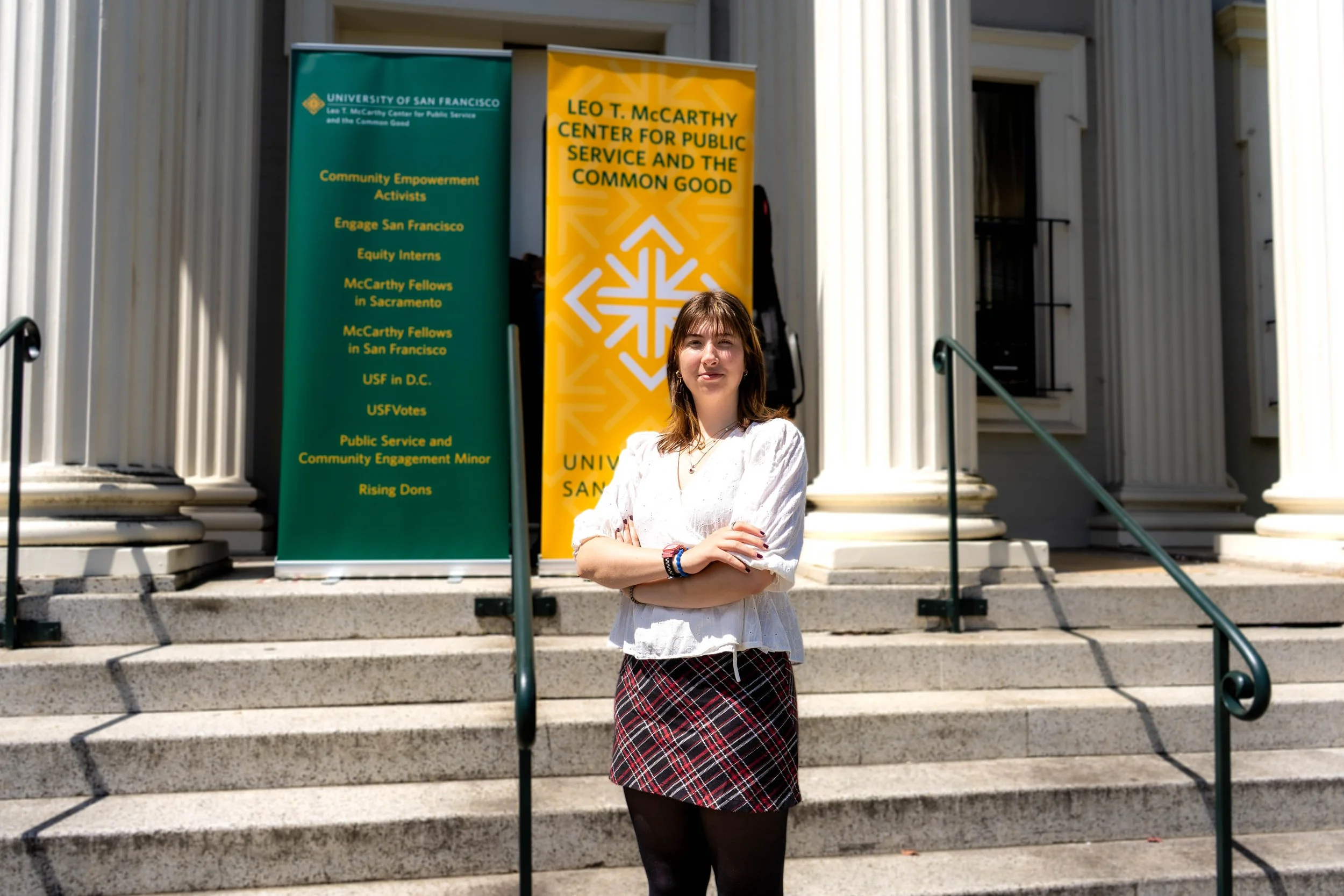 A young woman standing with arms crossed on the steps of a building, with two banners behind her indicating a university event at the University of San Francisco.