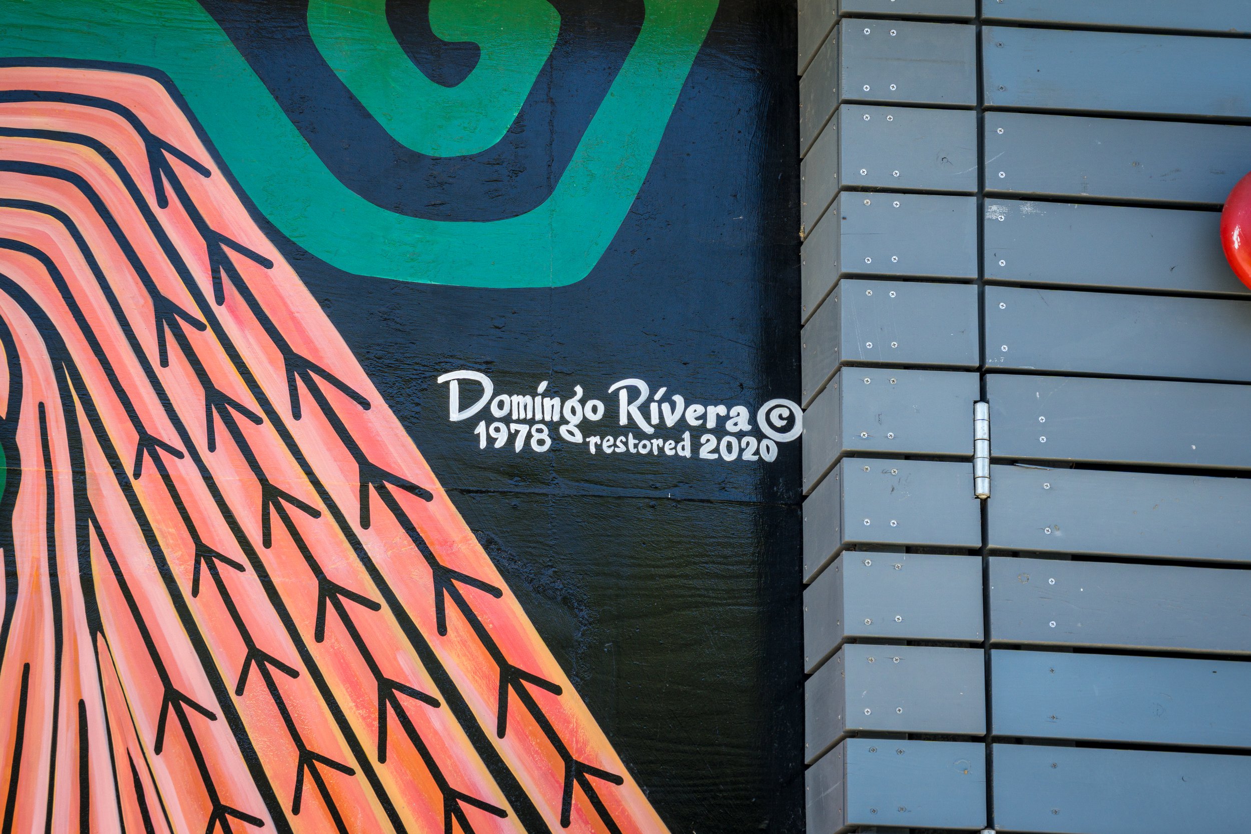 Mural art with colorful, abstract design on a dark background, signed by Domingo Rivera, with the dates 1978 and 2020 indicating restoration.