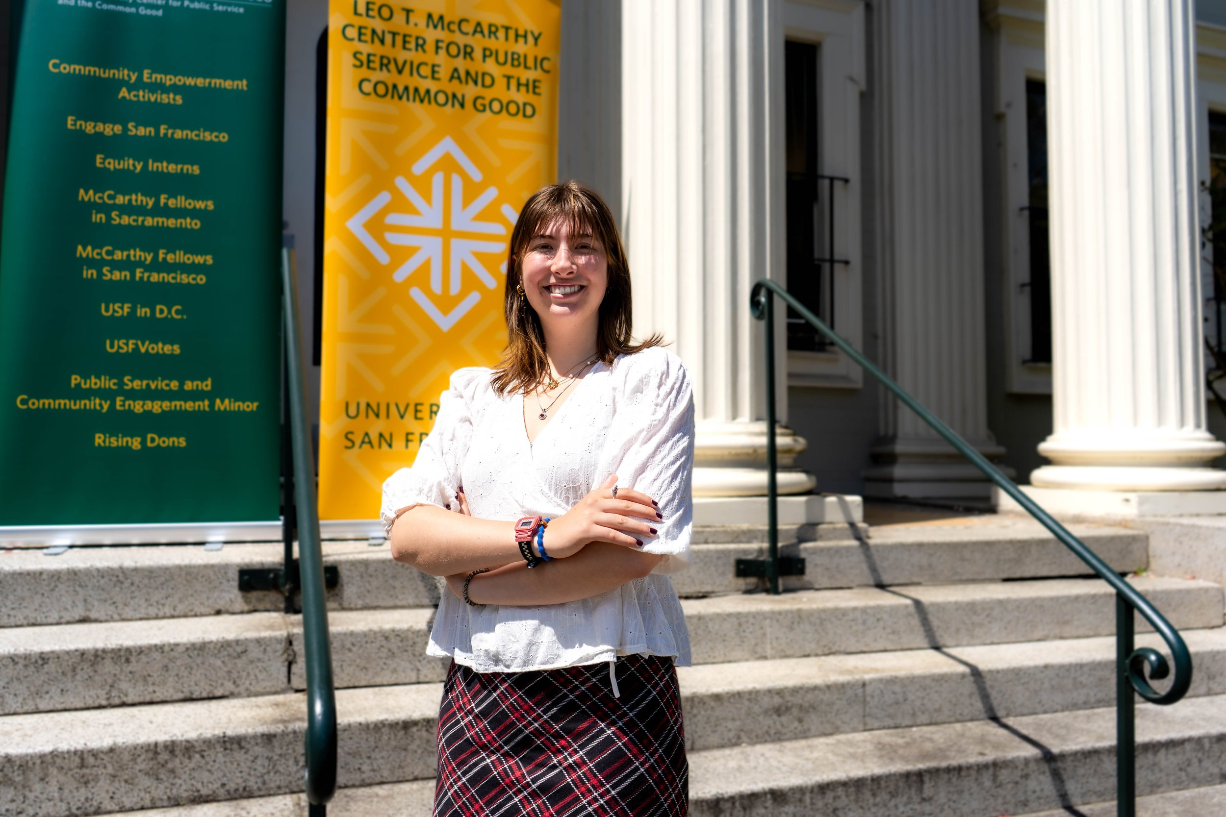 A young woman with shoulder-length brown hair standing on university steps, smiling, wearing a white blouse and plaid skirt, with banners and columns in the background.