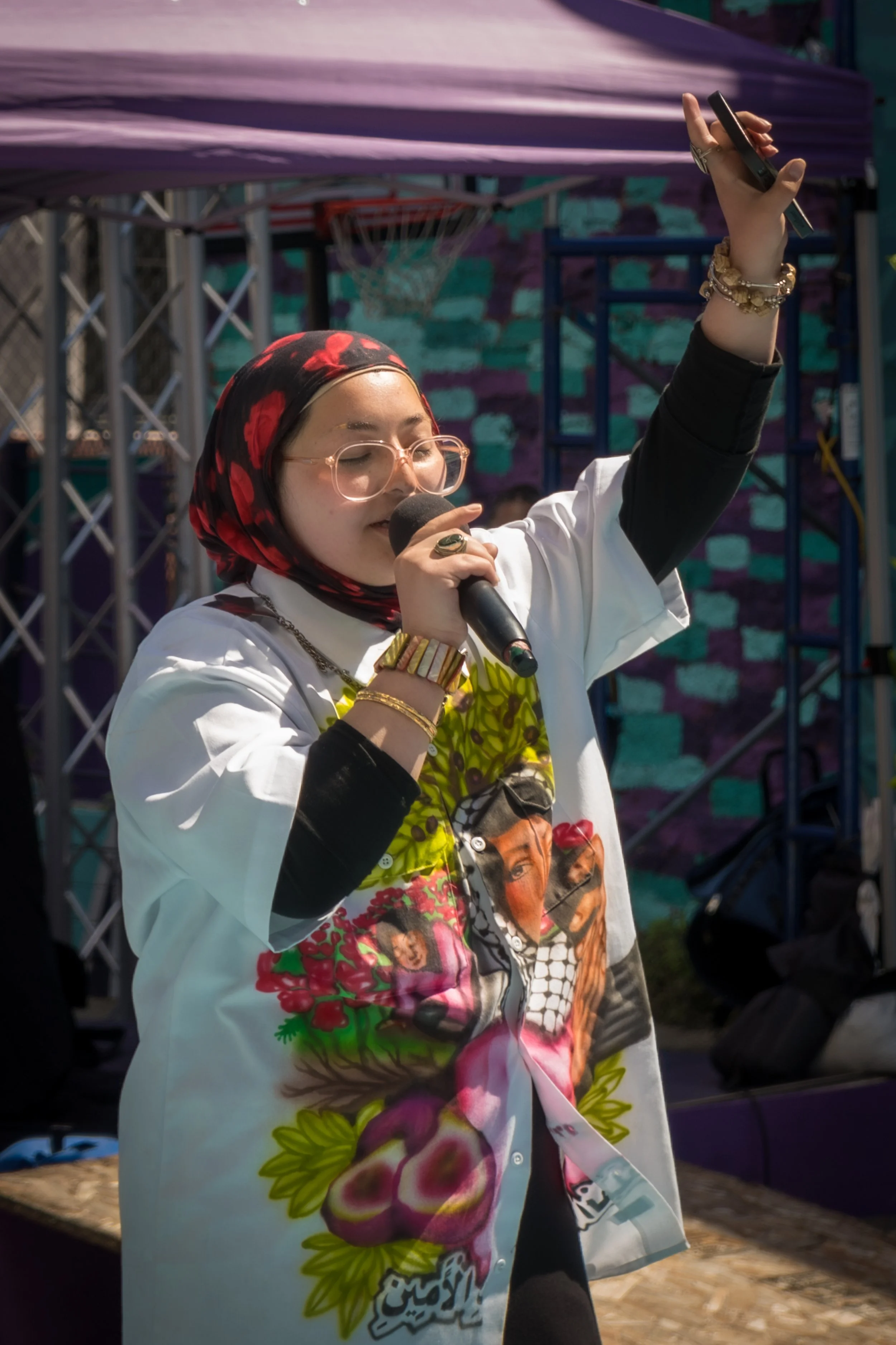 A woman wearing a colorful printed shirt, glasses, and a headscarf sings into a microphone with her eyes closed, holding a phone in her raised hand at an outdoor event.