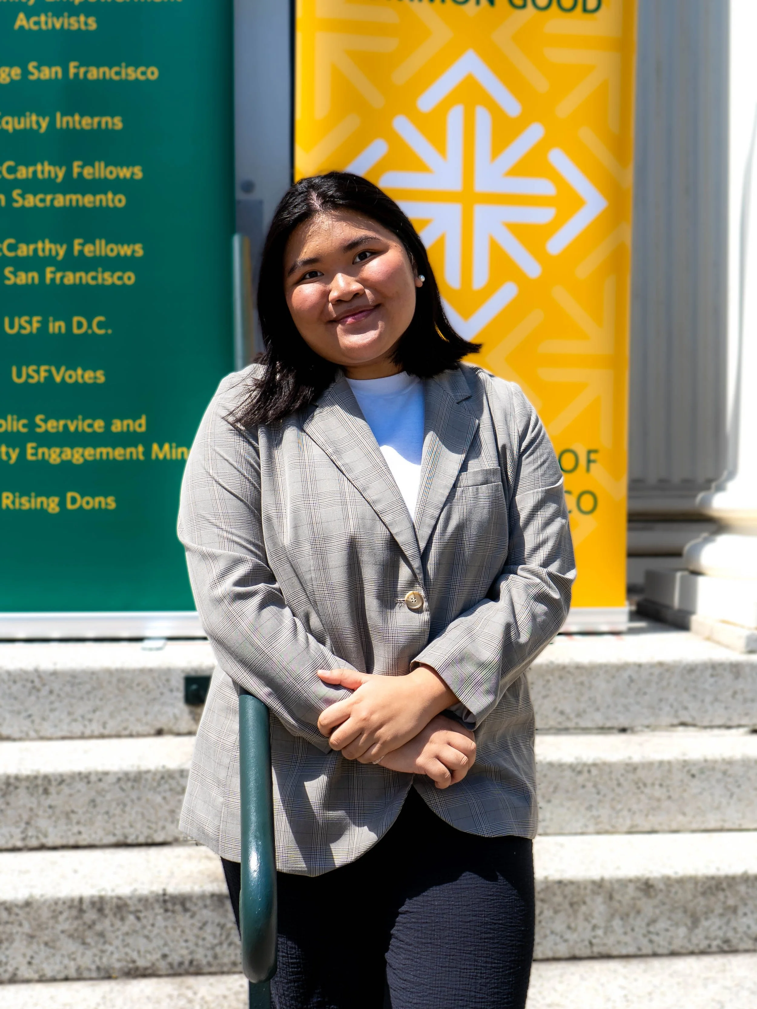 Young woman with shoulder-length black hair smiling, wearing a light plaid blazer and white shirt, standing on steps outdoors near a green and yellow informational sign.