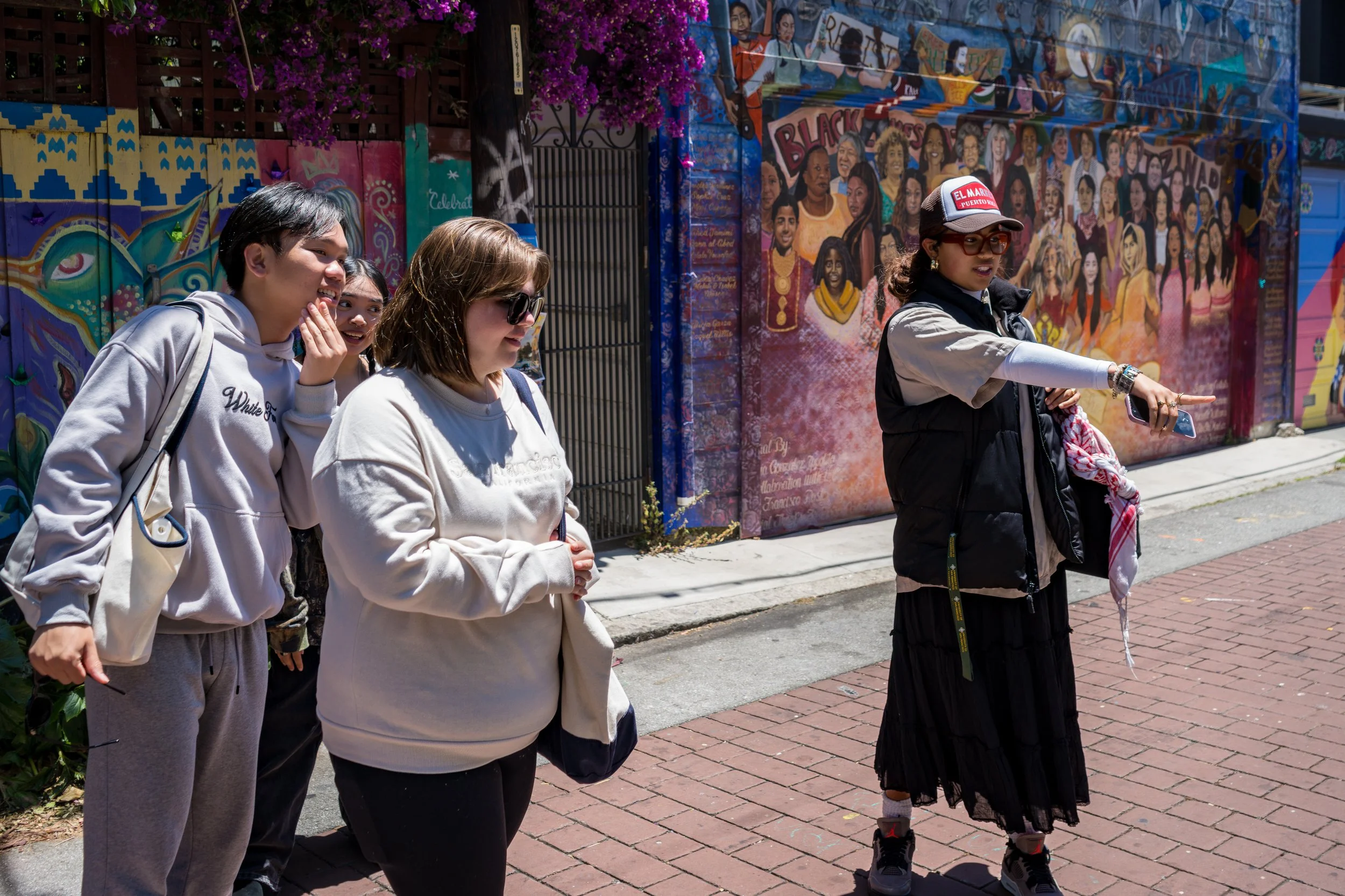 A tour guide pointing and talking to a group of four people on a city street with colorful murals in the background.