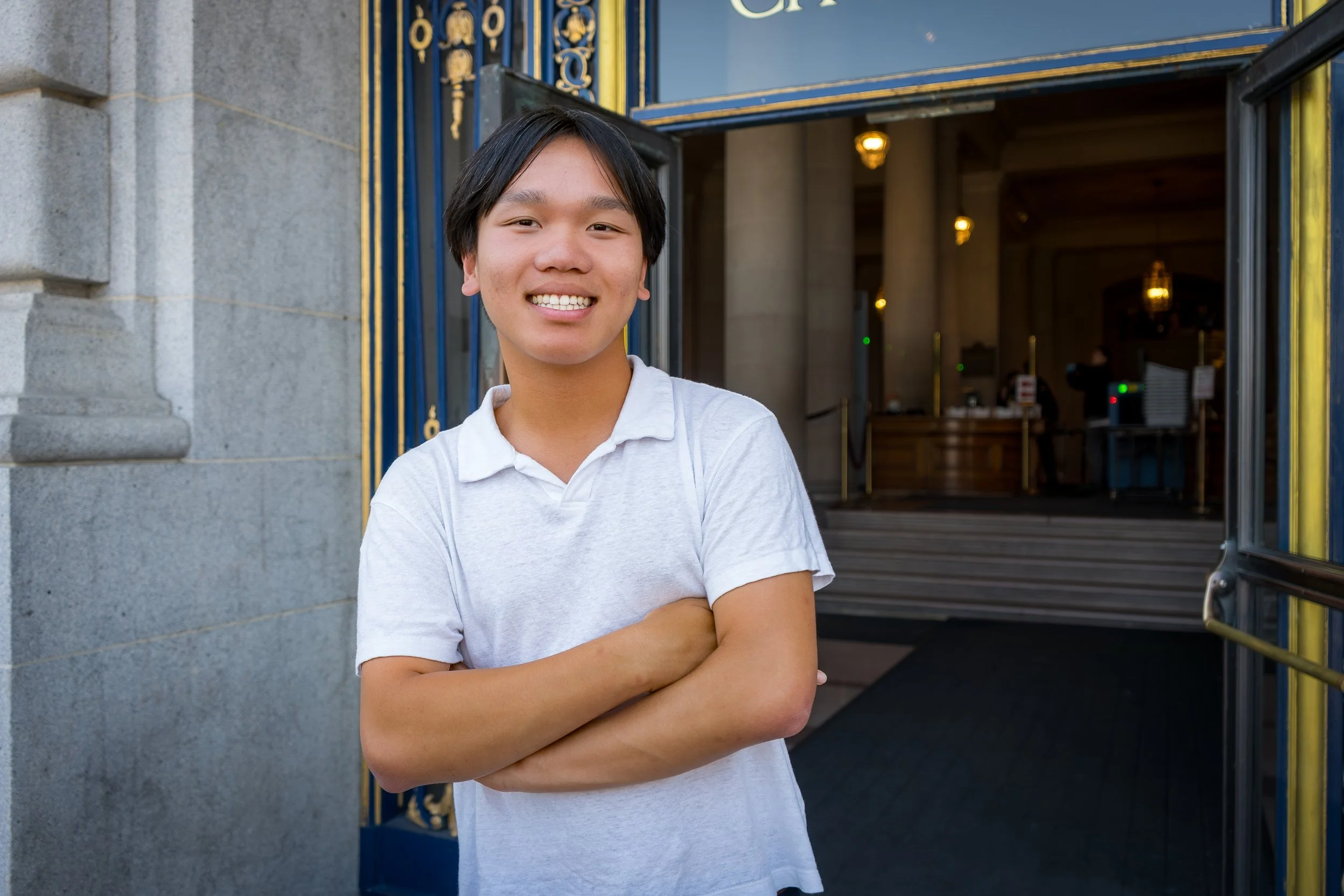 Young man with black hair in a white polo shirt smiling with arms crossed, standing outdoors in front of a building with ornate blue and gold gates.