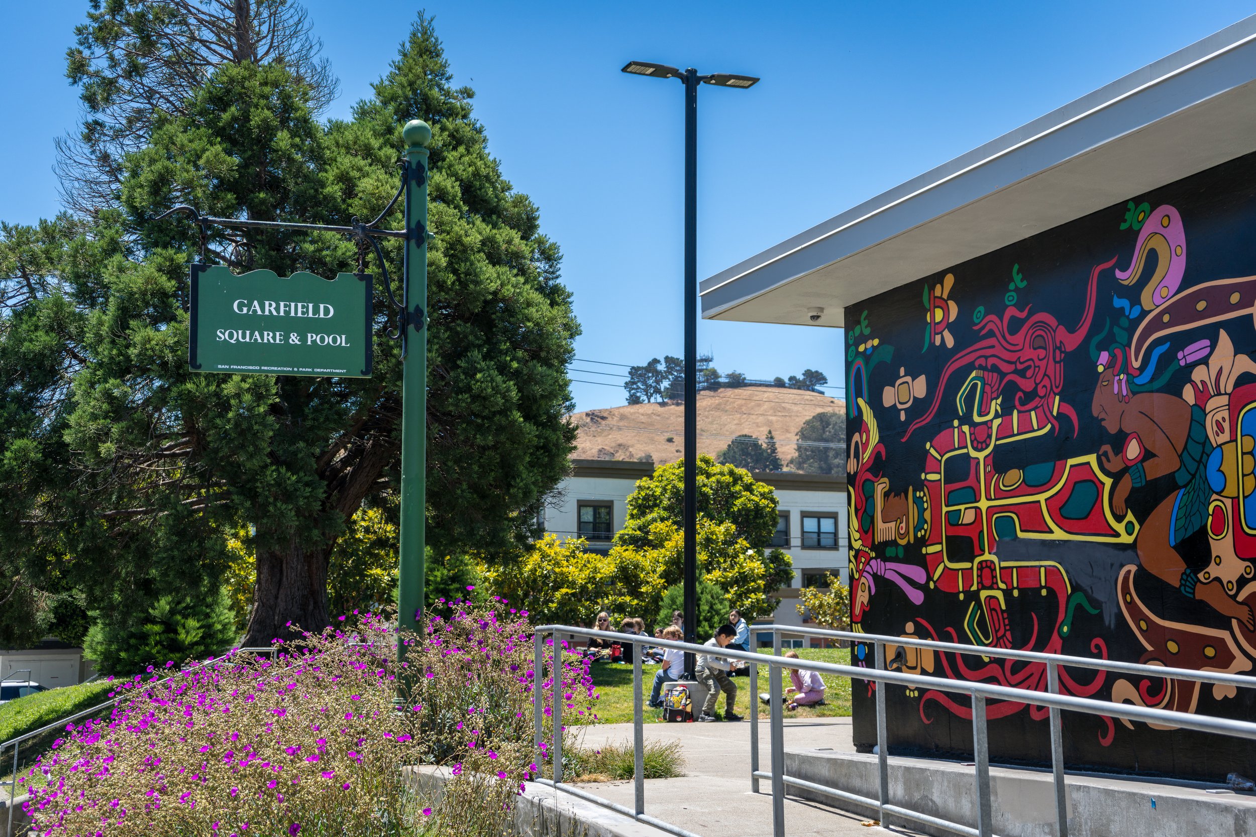 Street scene at Garfield Square & Pool with a mural on the right wall, a green sign, trees, flowers, and people sitting on grass in the background.