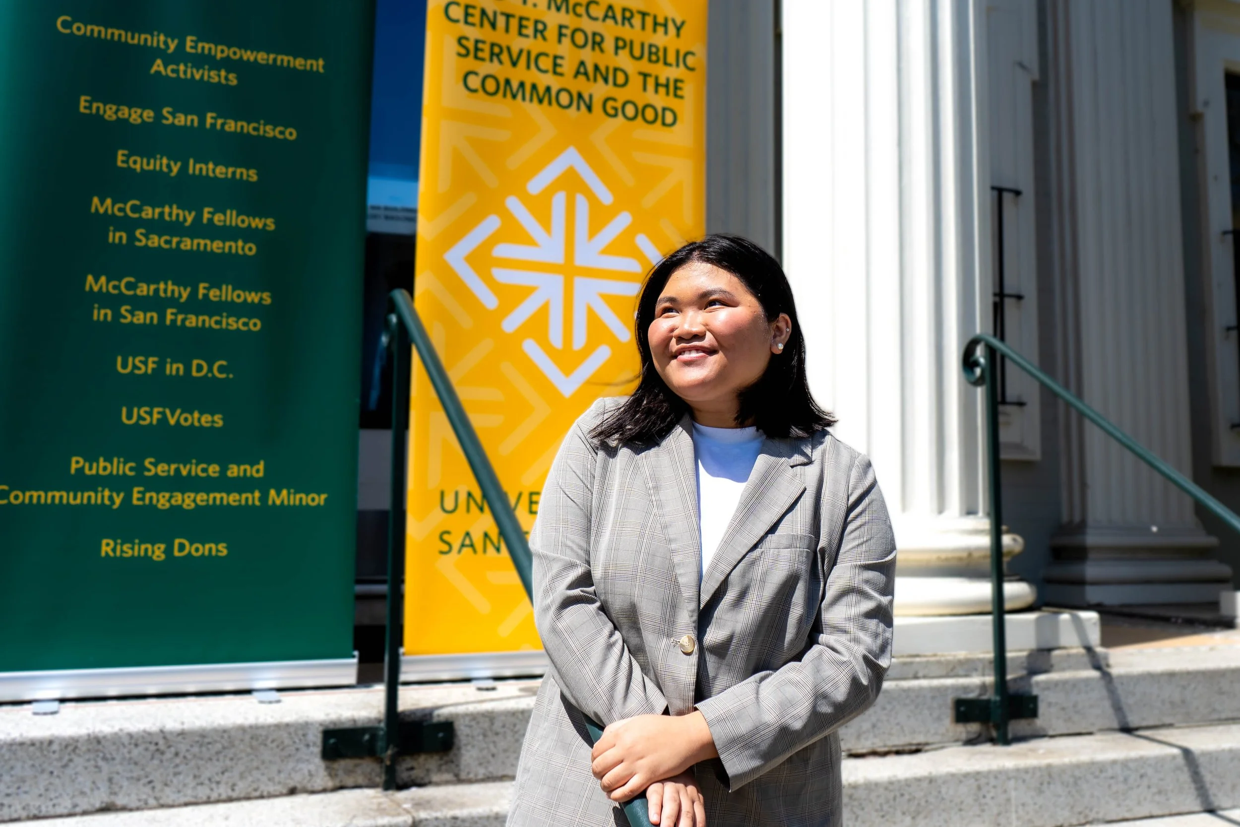 A woman in a plaid blazer standing outside a building with colorful signs in the background, one of which reads 'UNIVERSITY OF SAN FRANCISCO'.