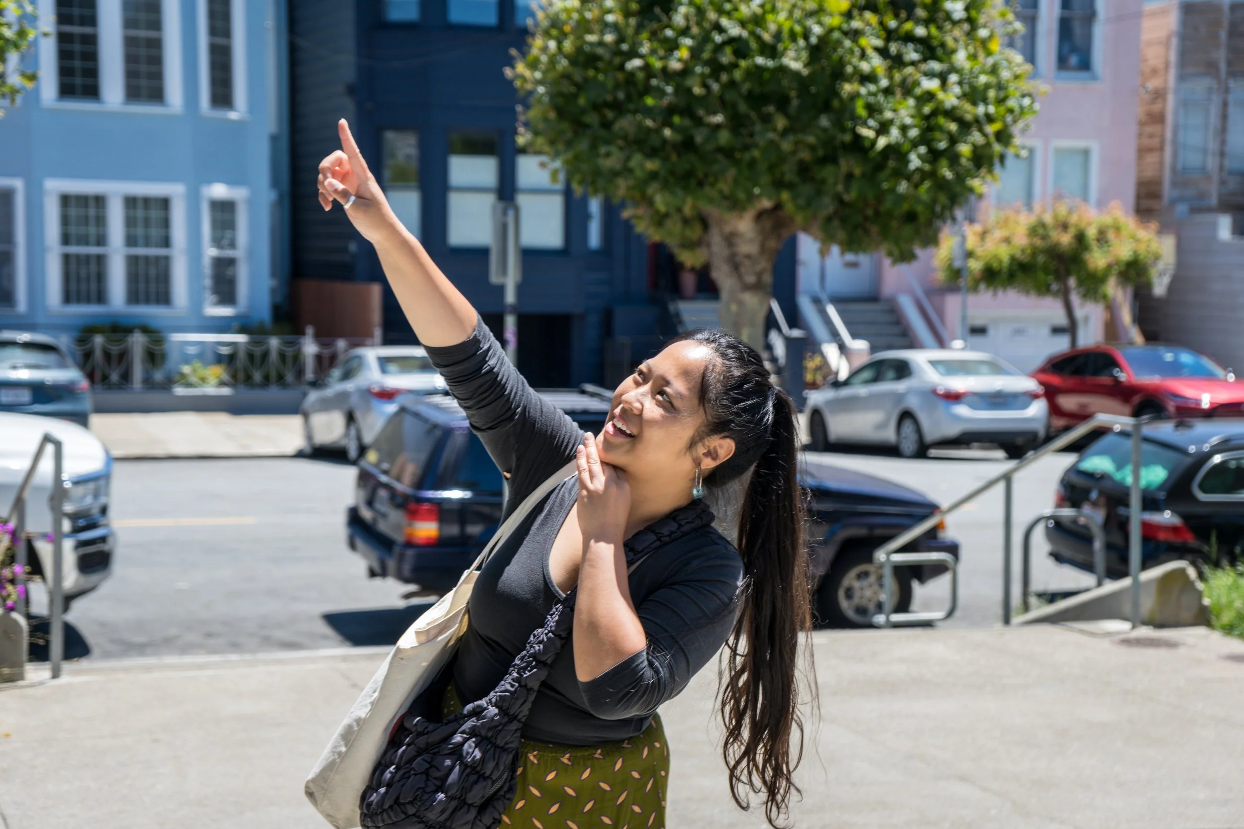 A woman standing outdoors on a sunny day, pointing up with her right hand while smiling and looking in the direction she is pointing. She has long dark hair, is wearing a black top, patterned pants, and has a beige tote bag on her shoulder.