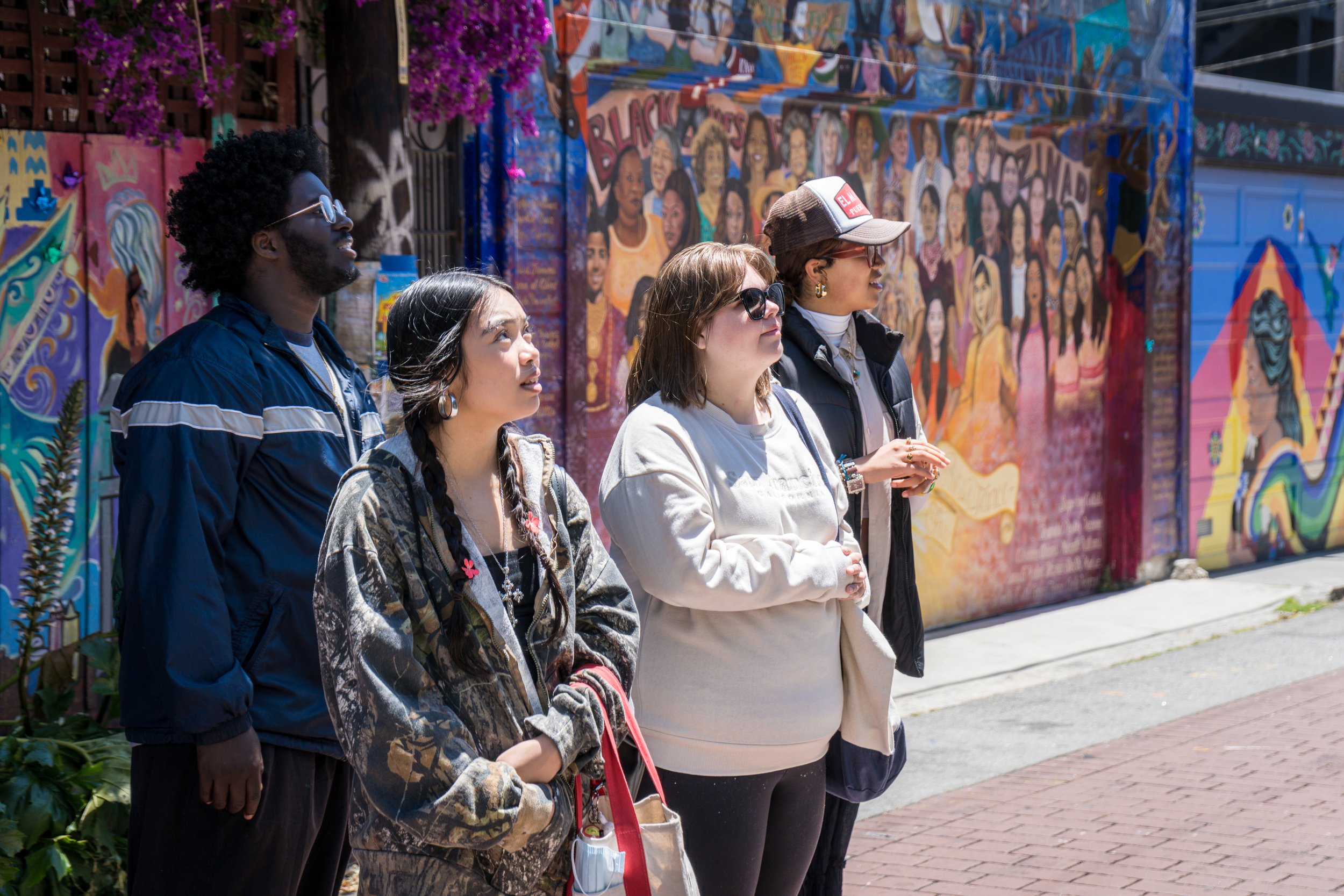 Four people standing outdoors near colorful mural art on a wall, looking in the same direction, enjoying a sunny day.