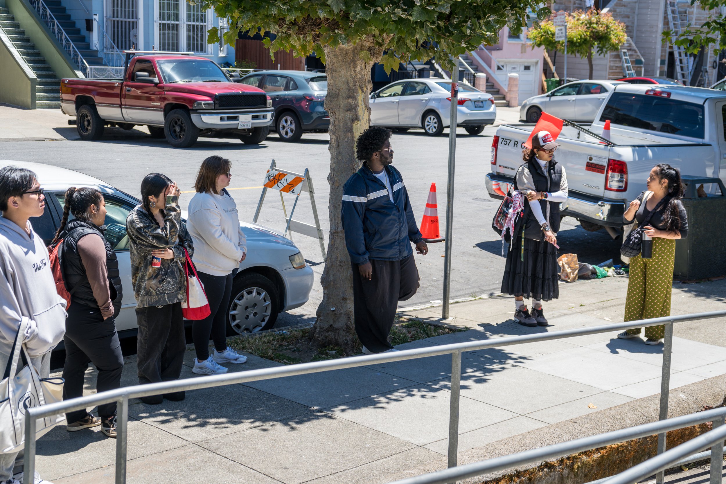 Group of people standing on the sidewalk, some wearing backpacks, in front of a row of parked cars and a large tree in an urban neighborhood with stairs and colorful houses in the background.
