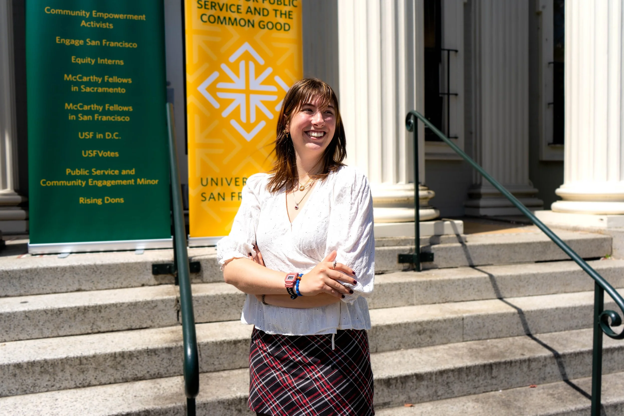 A young woman with shoulder-length brown hair and a white blouse stands on the steps outside a building with classical columns, smiling and crossing her arms. Behind her are two colorful banners with yellow text.
