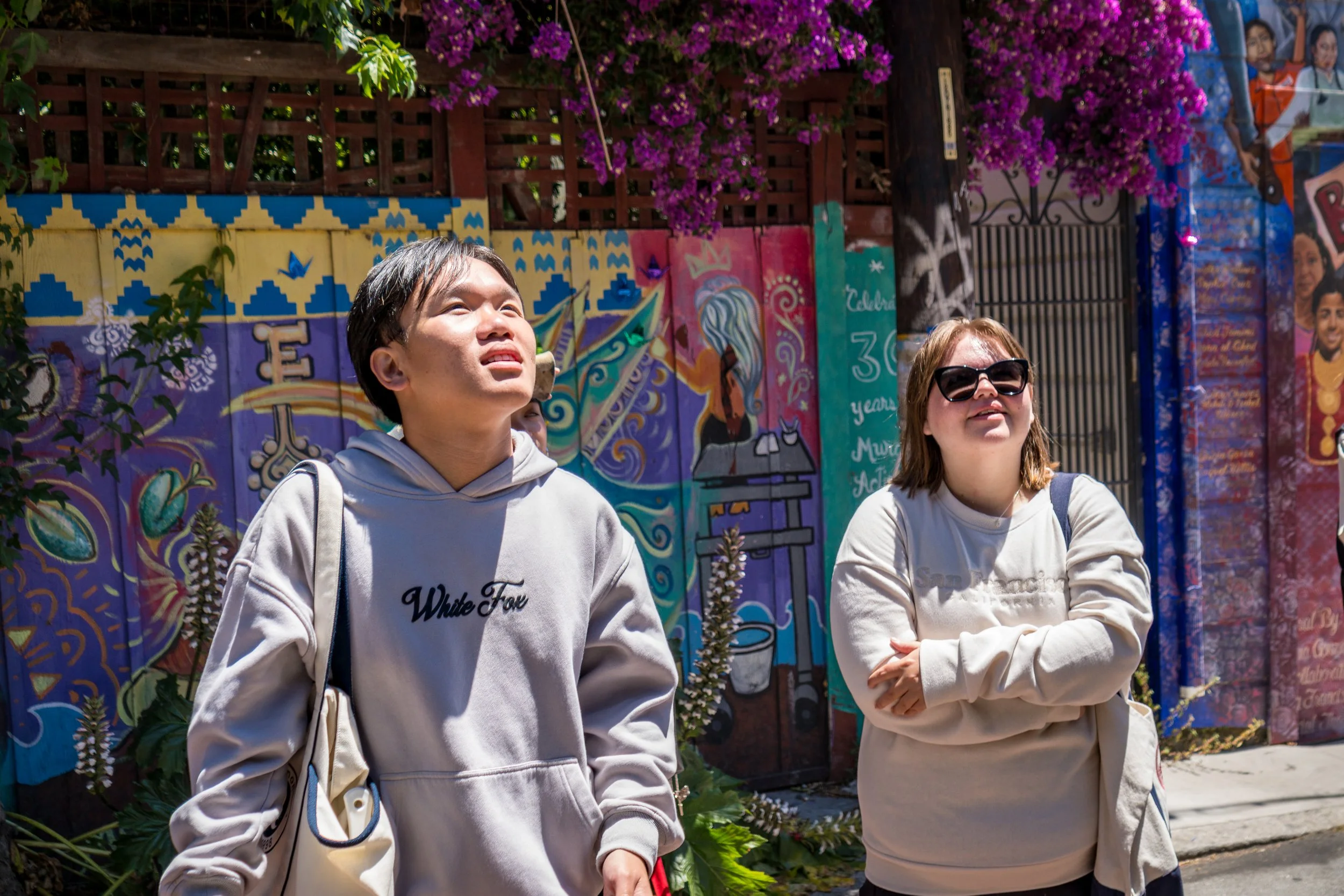 Two women standing outdoors in front of colorful murals and a flowering tree, looking up at something in the sky.