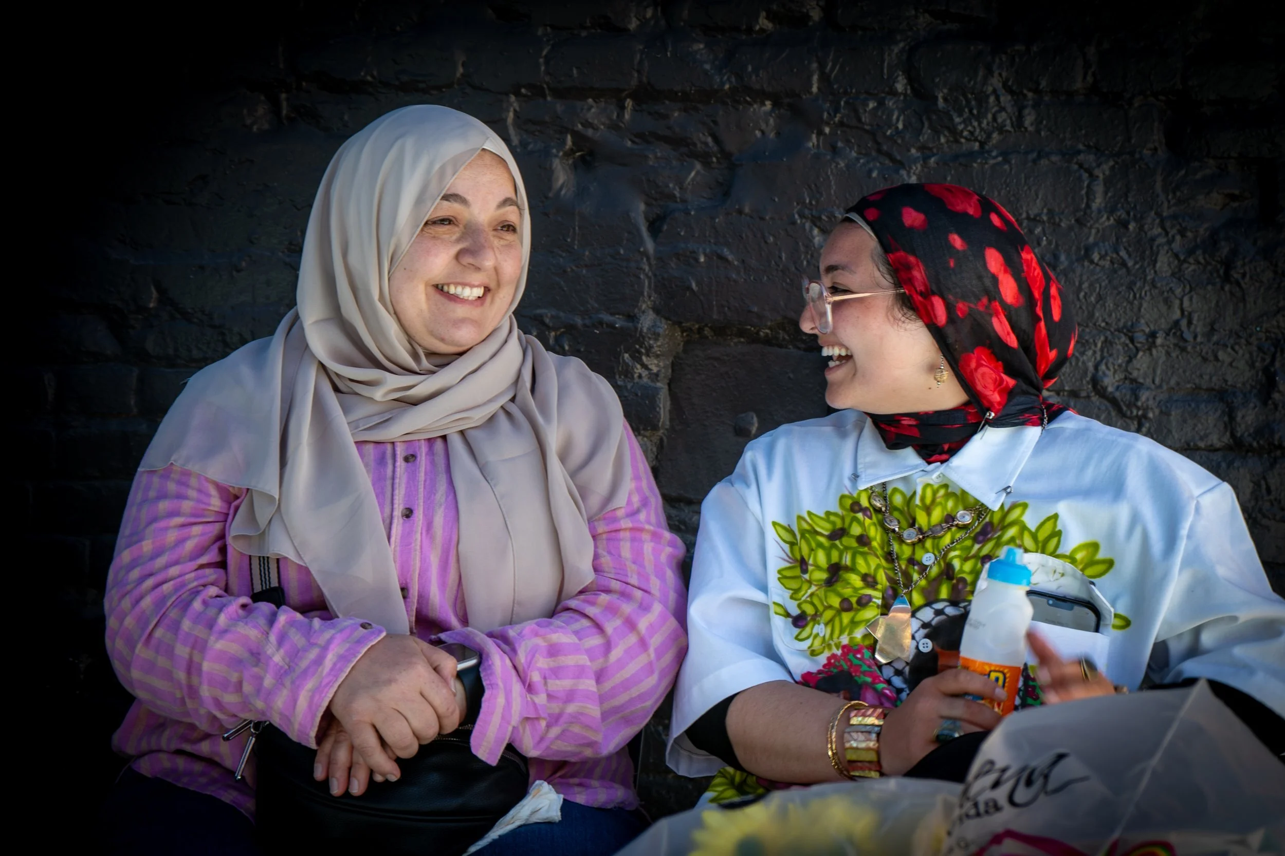 Two women wearing headscarves smiling and conversing in front of a dark stone wall.