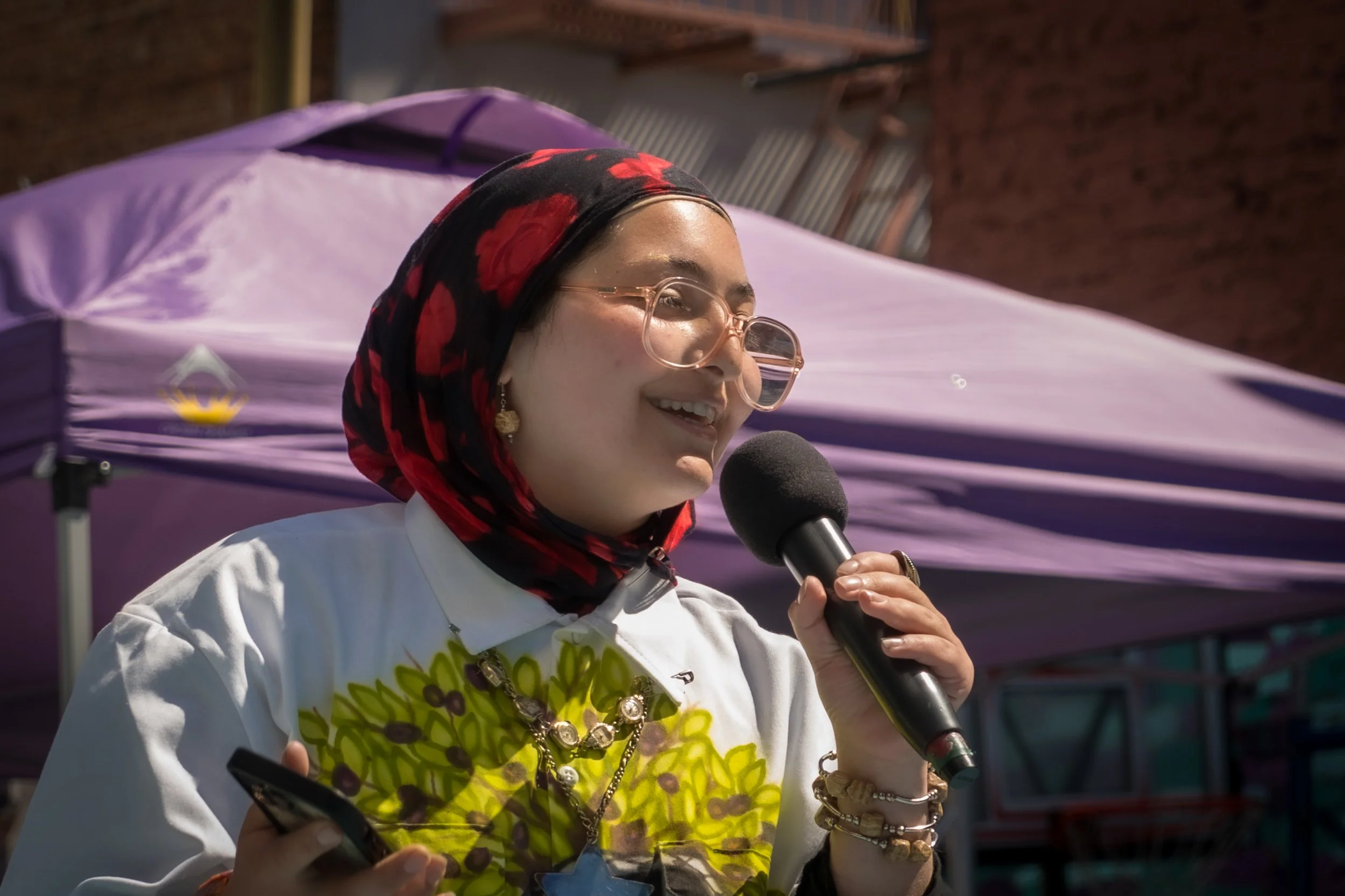 A woman with a colorful scarf, glasses, and jewelry smiling while speaking into a microphone outdoors with a purple canopy in the background.