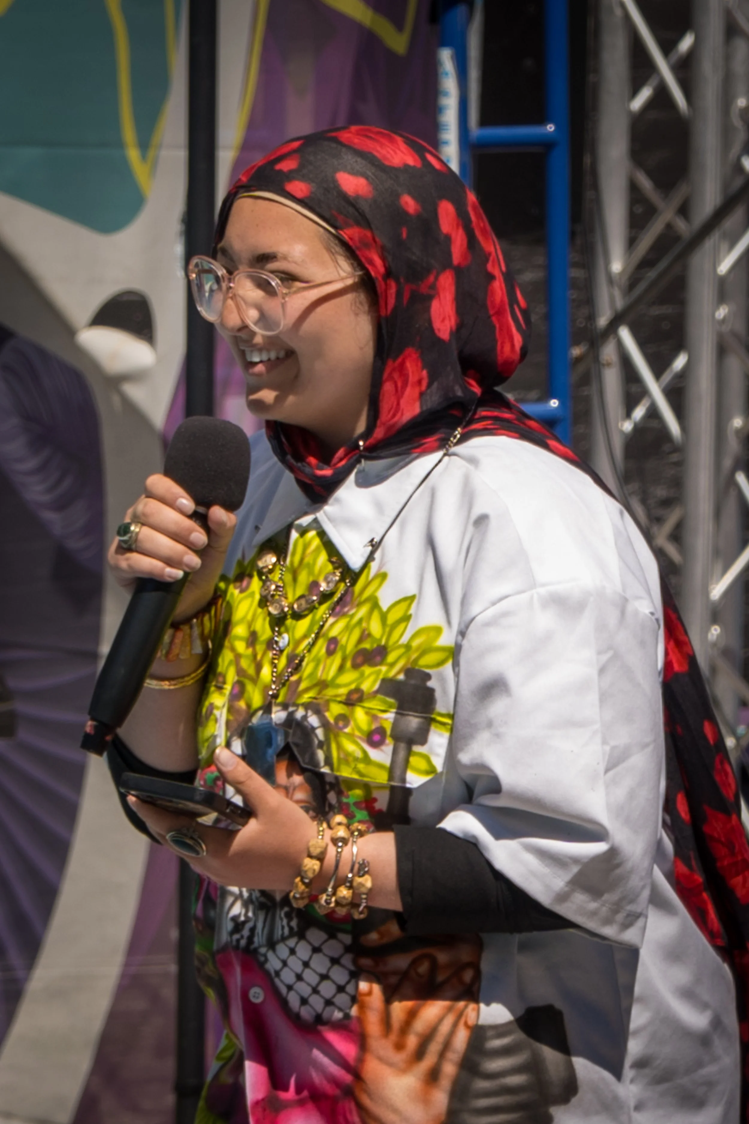 A woman wearing glasses, a red and black floral headscarf, and a colorful shirt, smiling while holding a microphone and a phone.