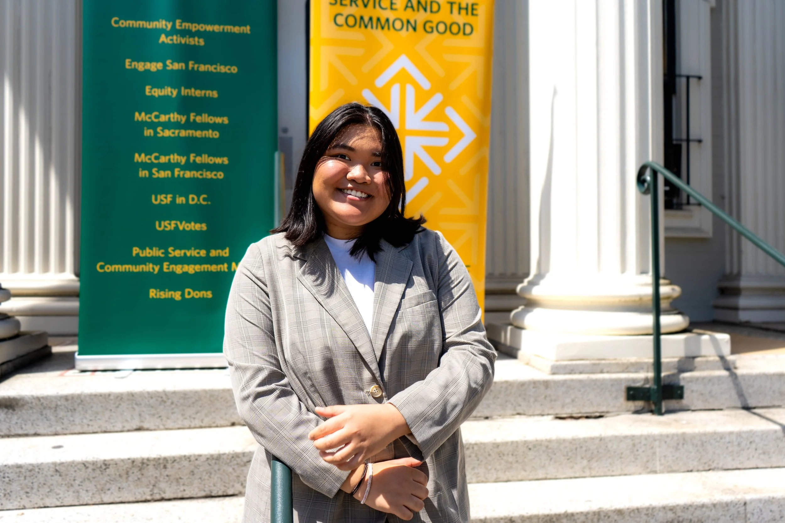 Young woman standing on steps outside a building, smiling, wearing a blazer, with banners behind her listing community programs and initiatives.
