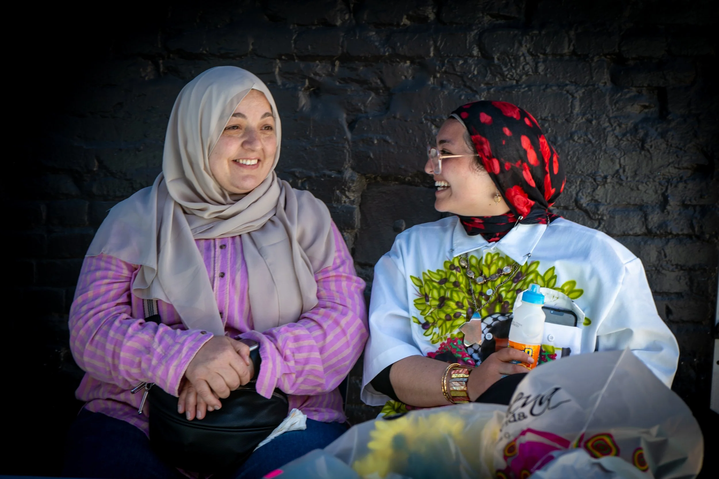 Two women sitting together, smiling and talking. The woman on the left is wearing a beige headscarf and a pink striped shirt. The woman on the right has a black headscarf with red flowers, glasses, and a white embroidered shirt. They are seated again