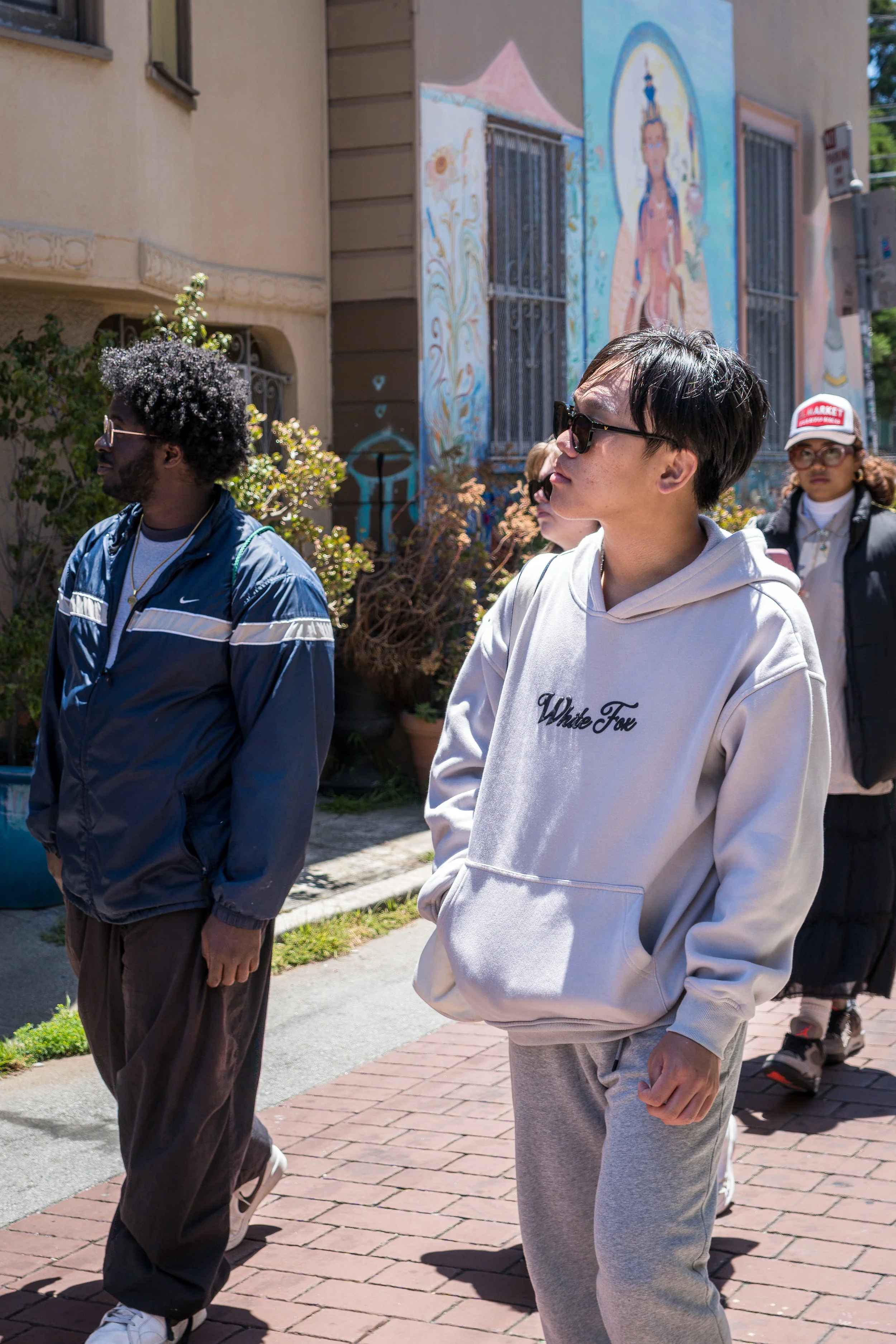 A group of diverse people walking outdoors on a sunny day, with a colorful mural on a building wall in the background.