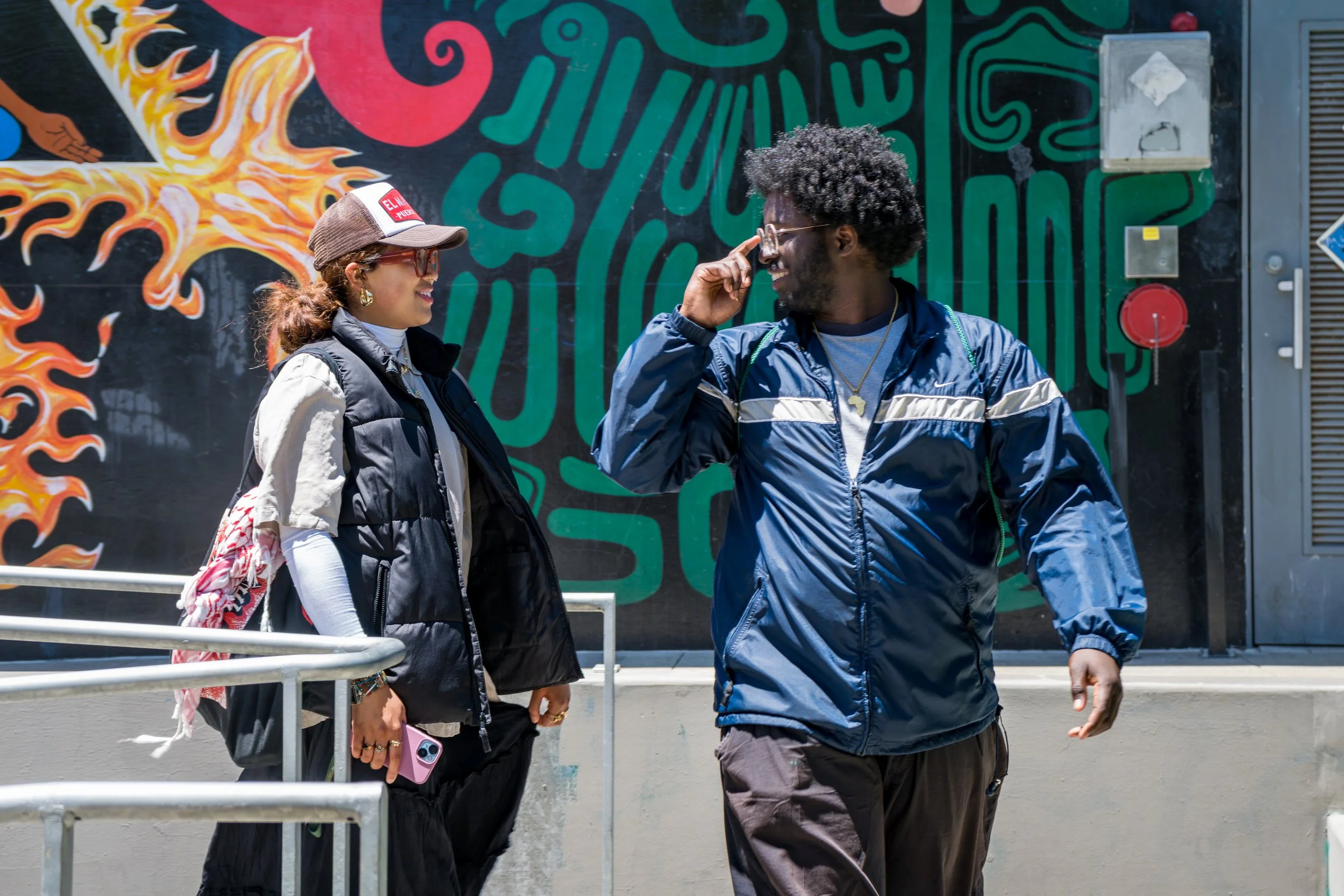 A man and woman are talking and smiling outdoors in front of a colorful mural. The woman wears a baseball cap, sunglasses, a black puffer vest over a beige and white shirt, and holds a pink phone. The man wears glasses, a blue windbreaker, and has an
