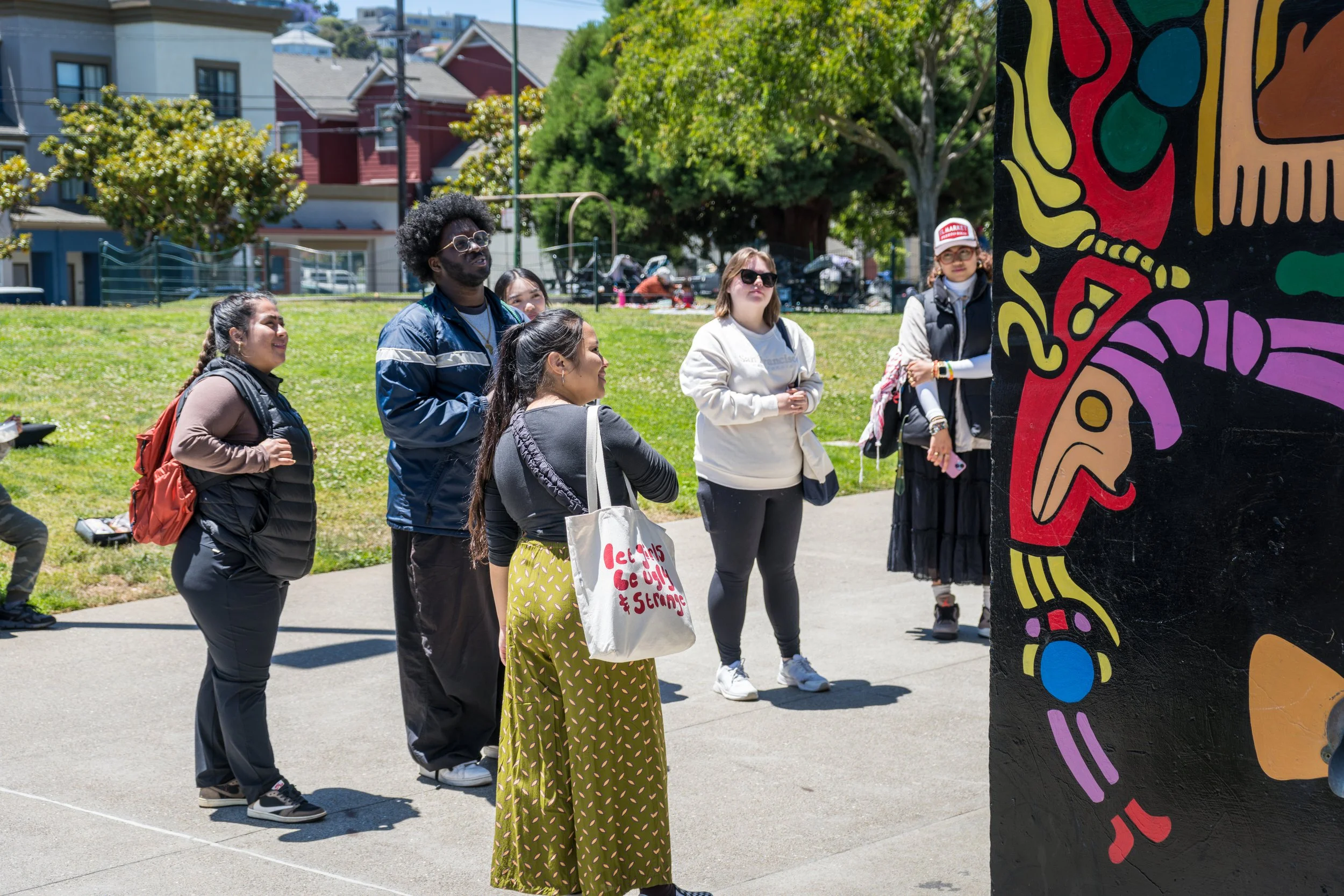 Group of diverse people standing outdoors, observing a colorful mural on a black wall during daytime.