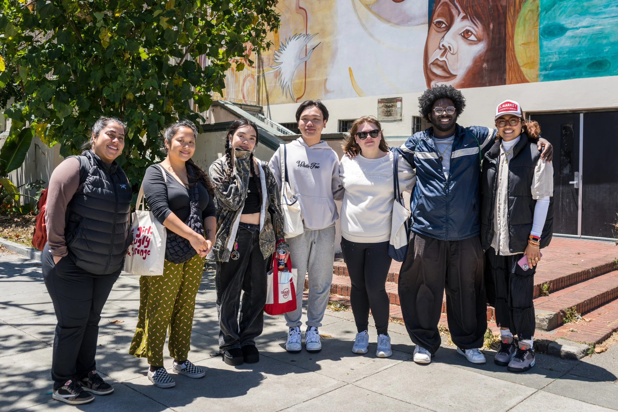 Group of eight diverse young people smiling and standing together outdoors in front of a mural wall. They are dressed casually and appear to be friends or classmates.
