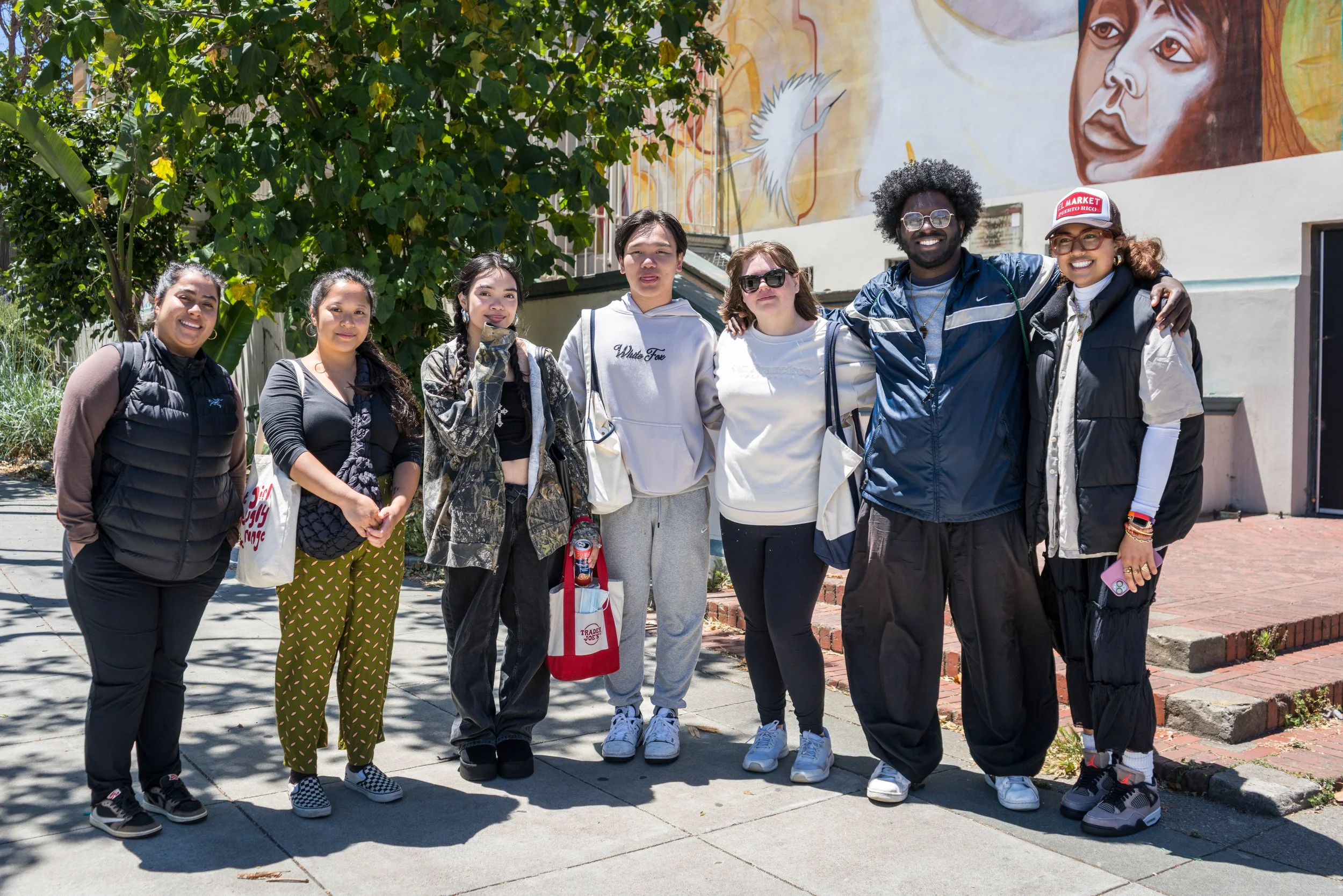 A diverse group of seven people standing together outdoors in front of a mural. They are smiling and dressed casually, with some wearing jackets and carrying bags.