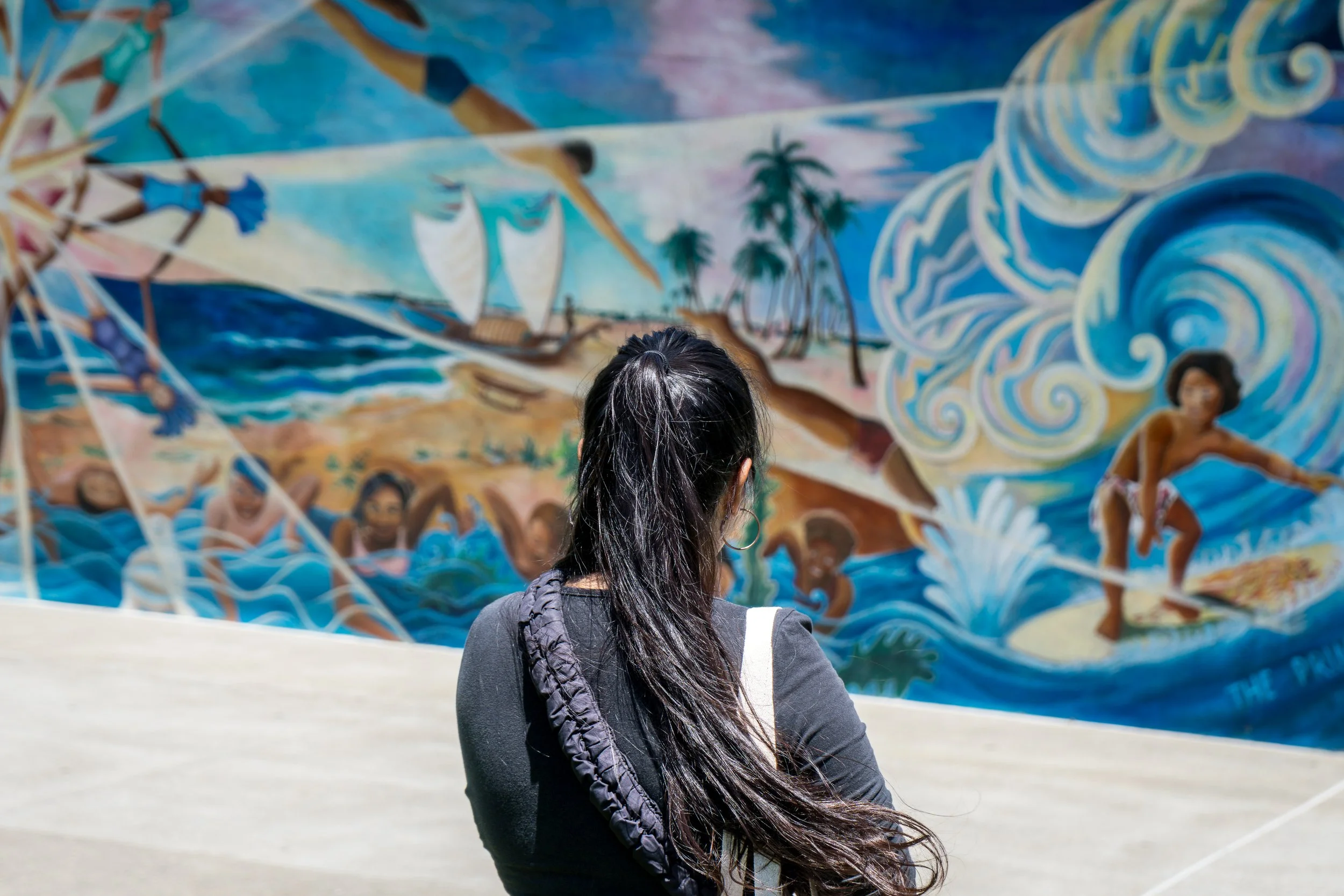 Woman with long black hair, wearing a black shirt, viewing a colorful mural depicting a pirate ship, ocean, palm trees, and children surfing on waves.