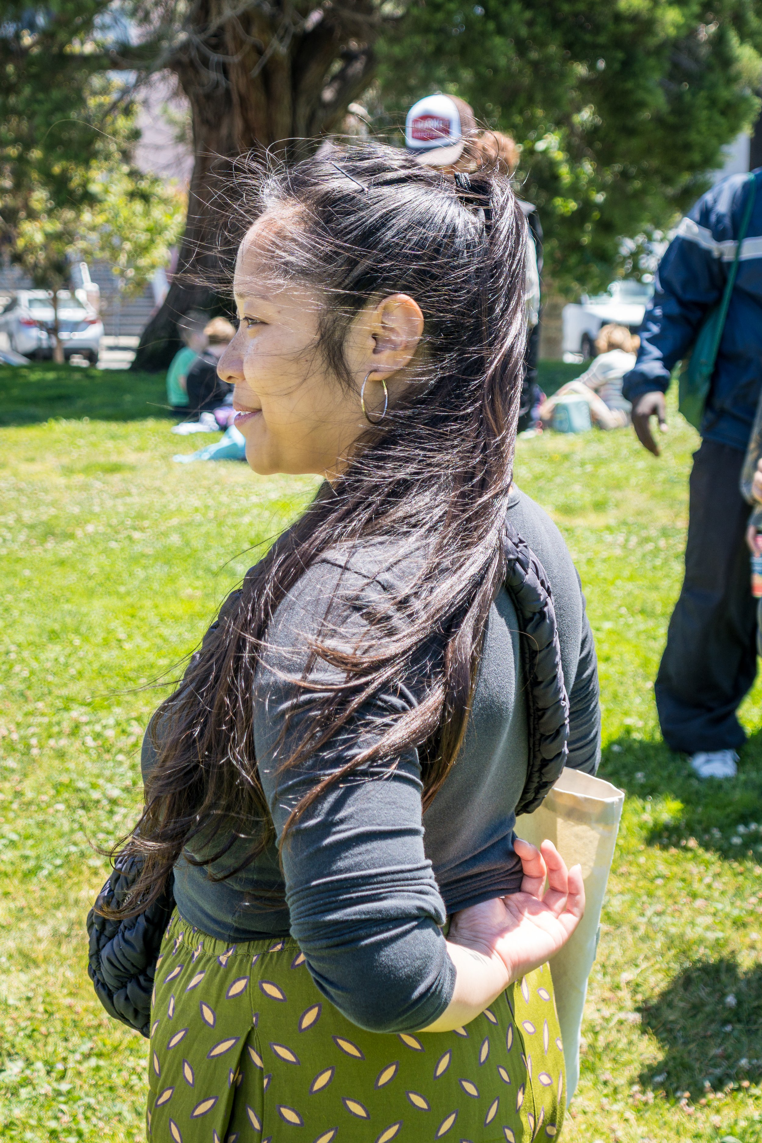 A woman with long dark hair, wearing a black top and a green skirt with a pattern, stands outdoors on a sunny day. She is smiling and looking to her left, with trees and other people in the background.