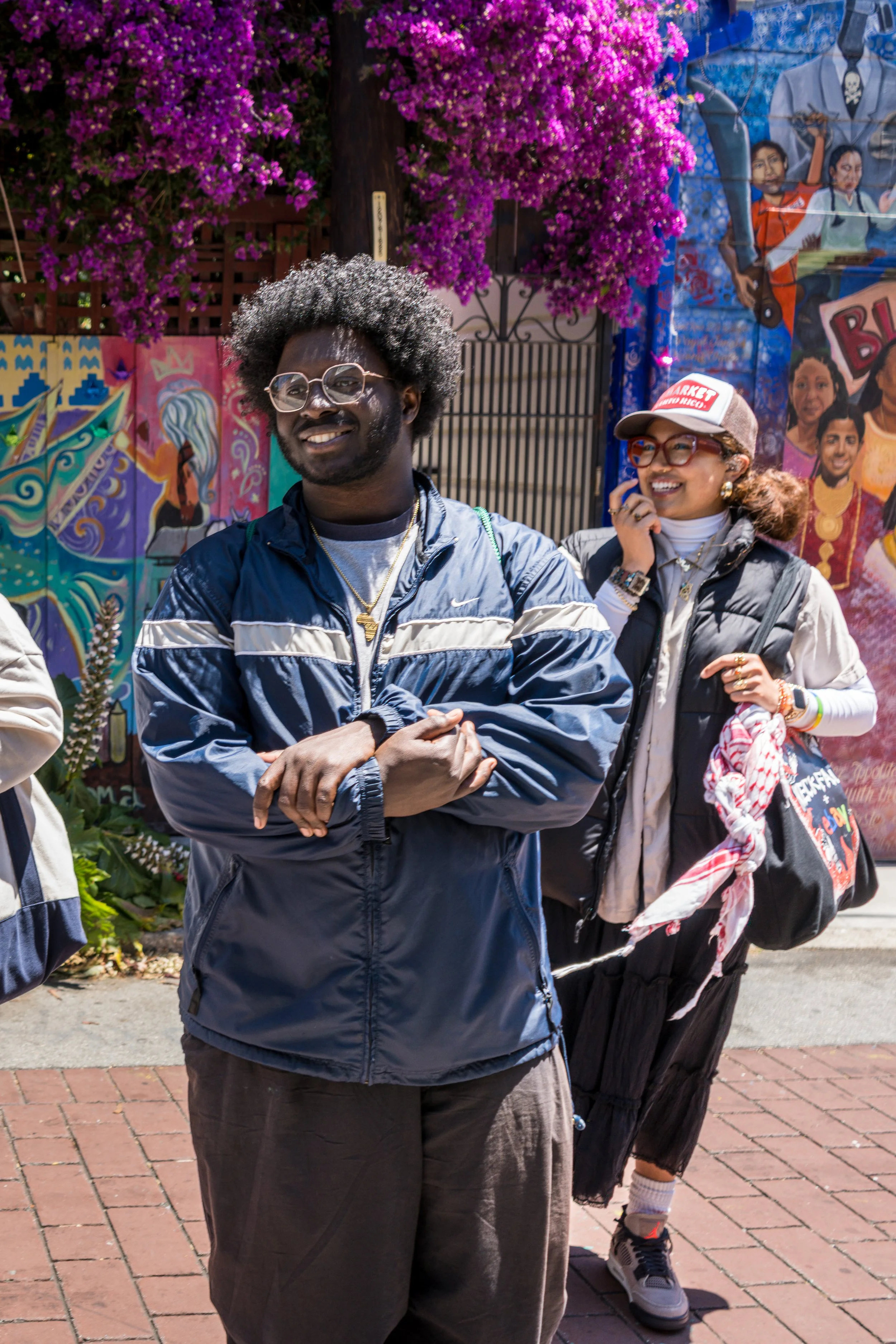 Two people smiling outdoors, standing in front of a colorful mural and bright pink bougainvillea flowers. The man in the foreground has curly hair, glasses, and wears a navy blue windbreaker. The woman behind him wears a cap, glasses, and a vest, hol
