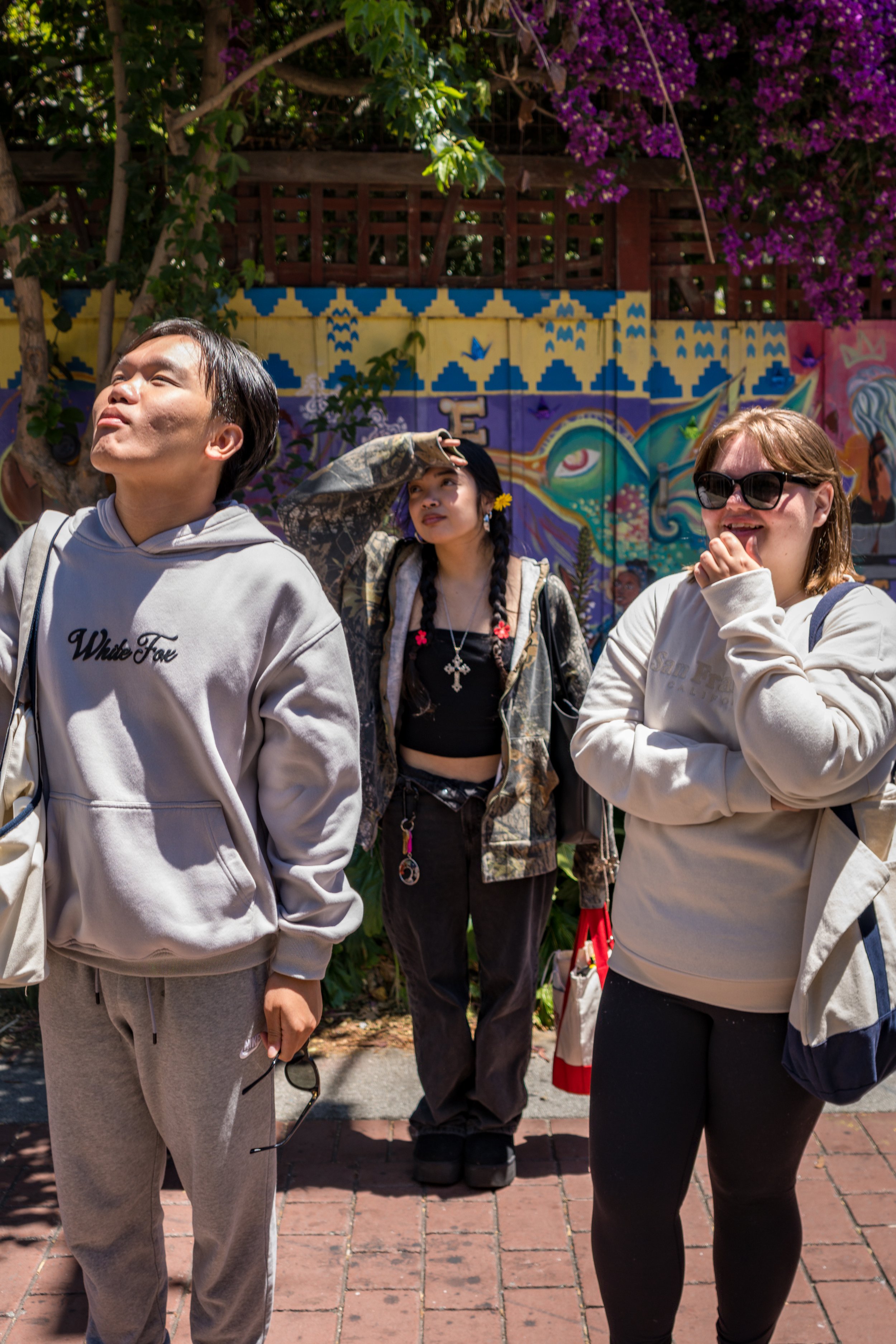 Three casually dressed young people standing outdoors near a colorful mural, with two of them looking up and one adjusting their hat, under a tree with purple flowers.