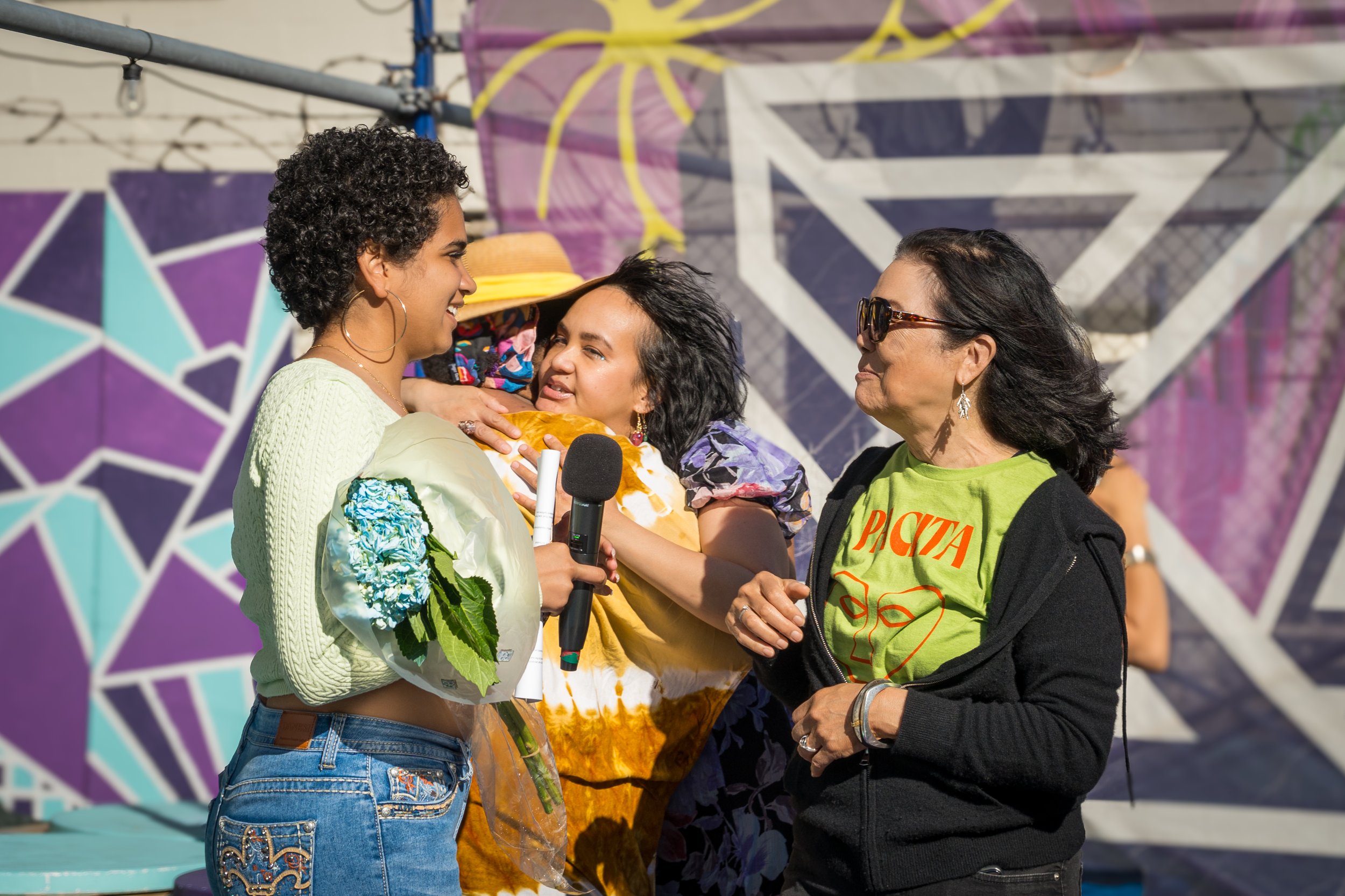 Three women interacting at an outdoor event with colorful geometric murals in the background. One woman holds flowers and a microphone, another woman is hugging her, and the third woman is observing, wearing sunglasses and casual attire.
