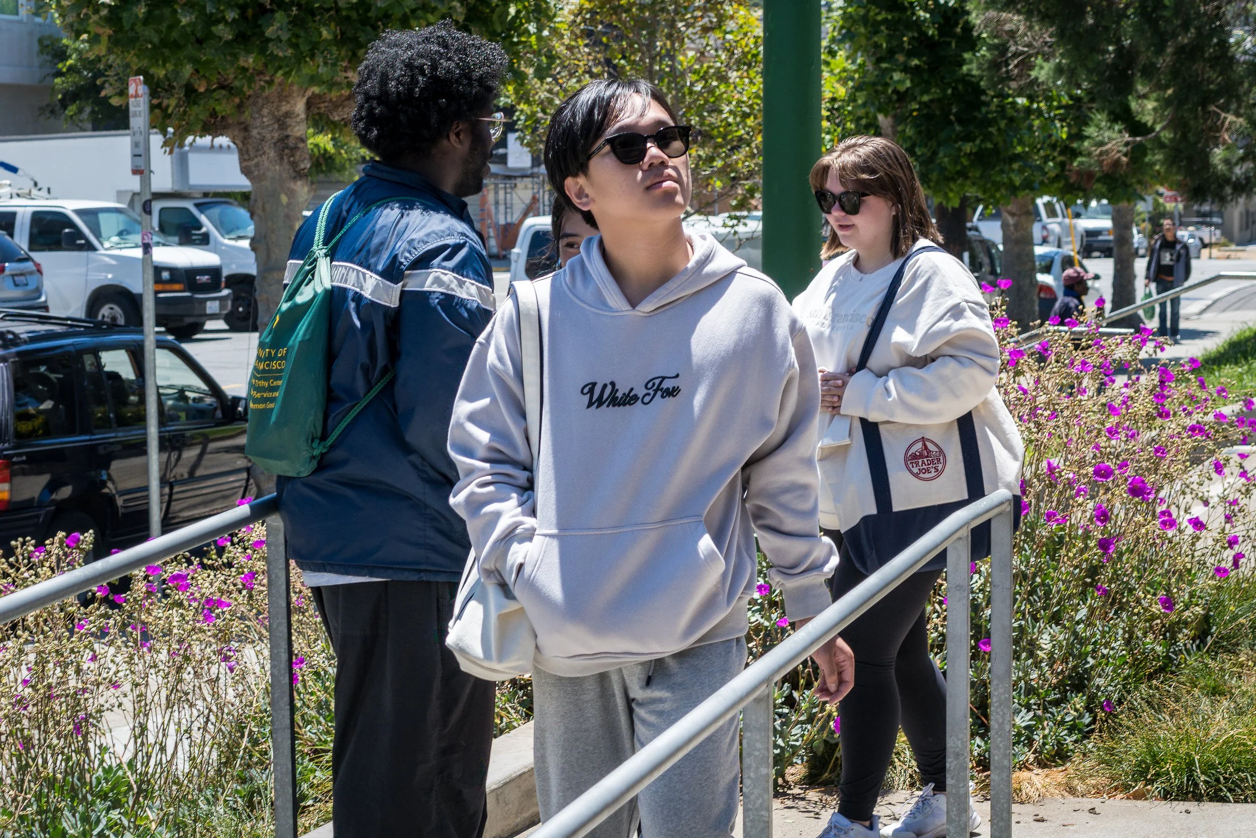 Group of four young adults, two women and two men, standing outdoors near a sidewalk with pink flowers, some wearing sunglasses and casual clothing including hoodies, jackets, and backpacks.