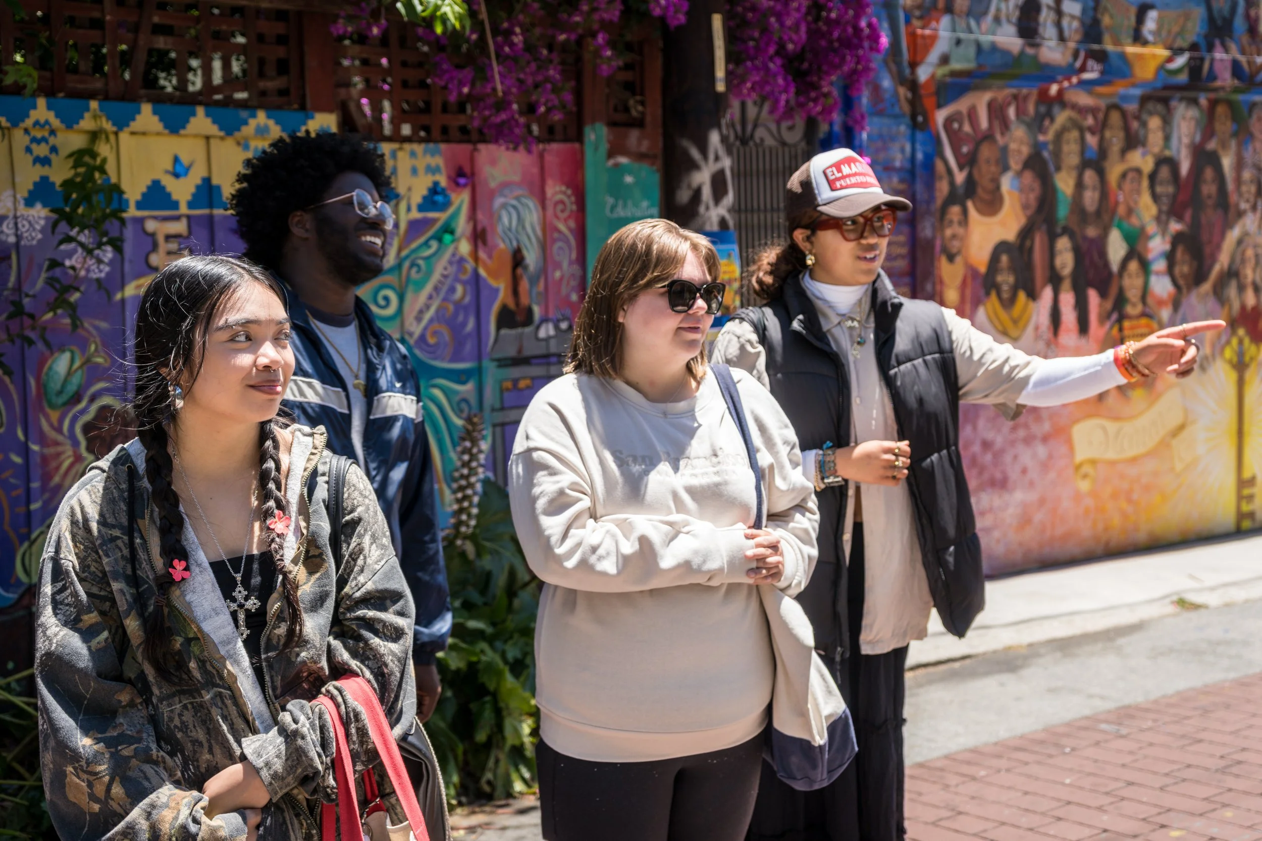A group of five diverse young women standing outdoors in front of a colorful mural, having a conversation.